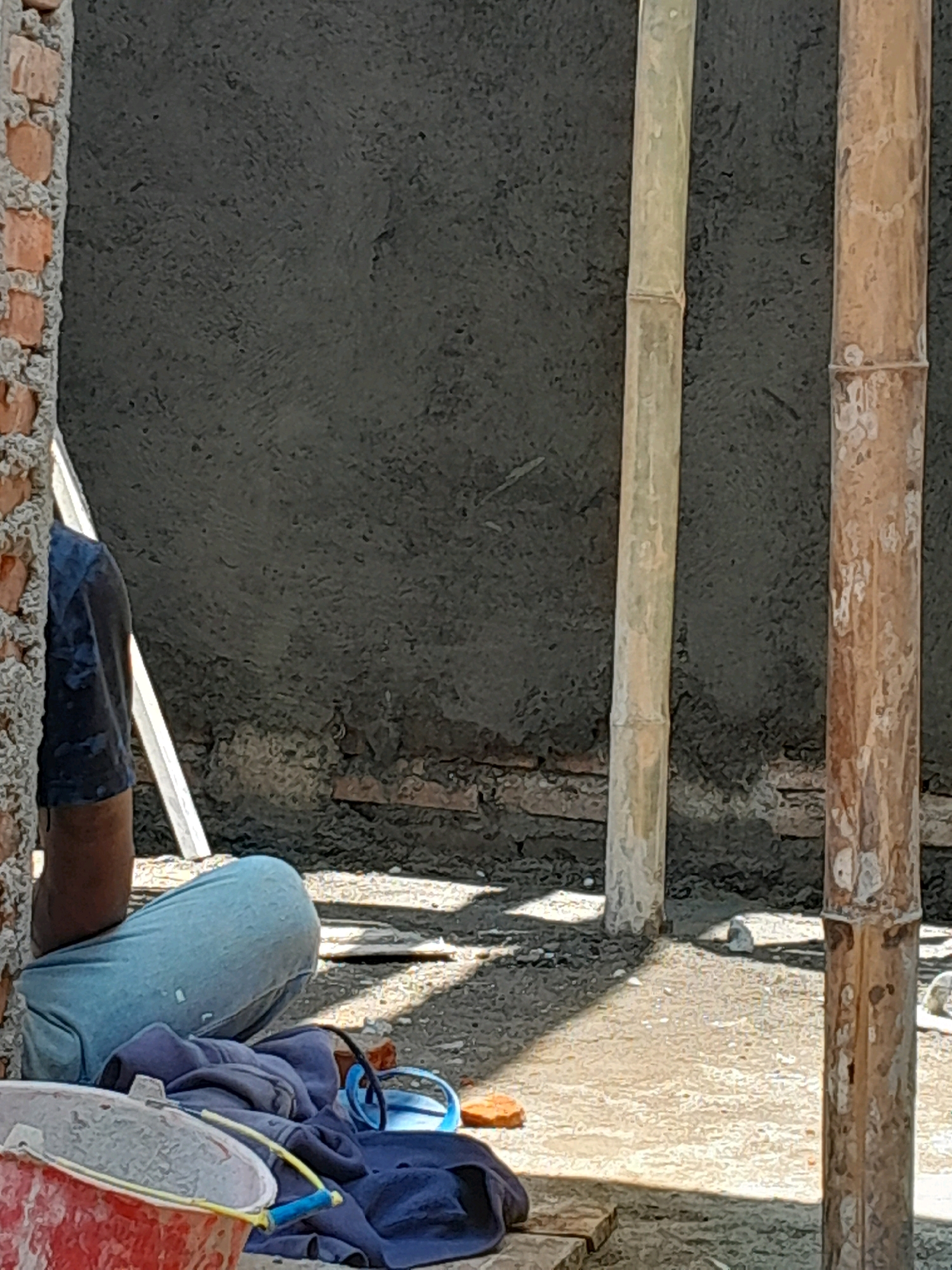 The image shows a construction site in Karangduren, Indonesia, with a worker in blue jeans and a dark blue t-shirt squatting on the ground. A red bucket is in the foreground, partially obscuring a pile of dark blue clothing and blue goggles. In the background, a rough, dark gray wall is visible, along with two wooden poles and a brick wall. The ground is covered with dirt and debris, and shadows are cast by the poles.