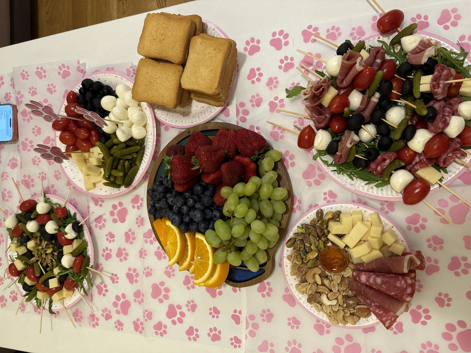 An overhead shot displays a table covered with a white cloth adorned with a repeating pattern of pink paw prints, indicating a themed gathering or party. The table is laden with various snack platters and appetizers.
In the upper right, a stack of golden-brown toasted bread slices rests on a white paper plate with a pink rim. Multiple white paper plates hold skewers featuring an assortment of ingredients, including cherry tomatoes, small mozzarella balls, black olives, small gherkins, and cubes of cheese, often arranged on a bed of arugula. Some skewers also include folded slices of prosciutto and salami.
A central wooden serving board is filled with fresh fruits: vibrant red strawberries, dark blue blueberries, clusters of green grapes, and bright orange slices. Another white paper plate contains a mix of cubed cheese, pistachios, cashews, and almonds, alongside folded slices of salami and prosciutto, with a small clear dish of honey. One platter of vegetables and mozzarella balls is decorated with pink paw-print picks.
A smartphone is partially visible on the left side of the table, resting on the paw-print tablecloth, but its screen content is illegible. The scene suggests an indoor, casual event focused on communal snacking. No people are visible.
