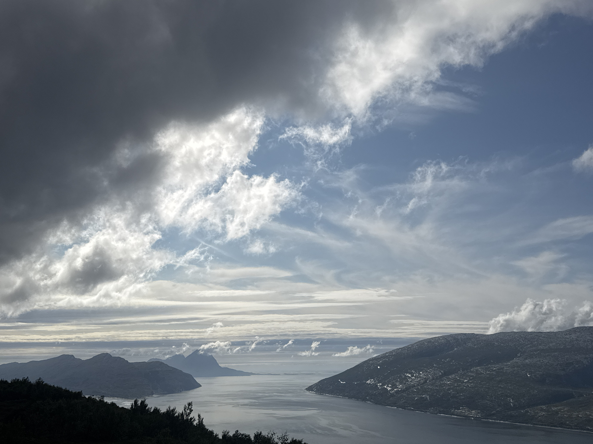 The image is a panoramic view of a fjord landscape in Flostrand, Norway. In the foreground, a dark, silhouetted treeline covers the bottom of the frame. Beyond this, a body of calm, grey water, likely a fjord, stretches into the distance. Several mountainous islands, shrouded in varying degrees of shadow and light, rise from the water. The sky is dramatic, with a mix of dark, heavy clouds on the left and lighter, wispy clouds against a blue sky on the right. The overall scene suggests a cool, possibly windy day with changing weather conditions.