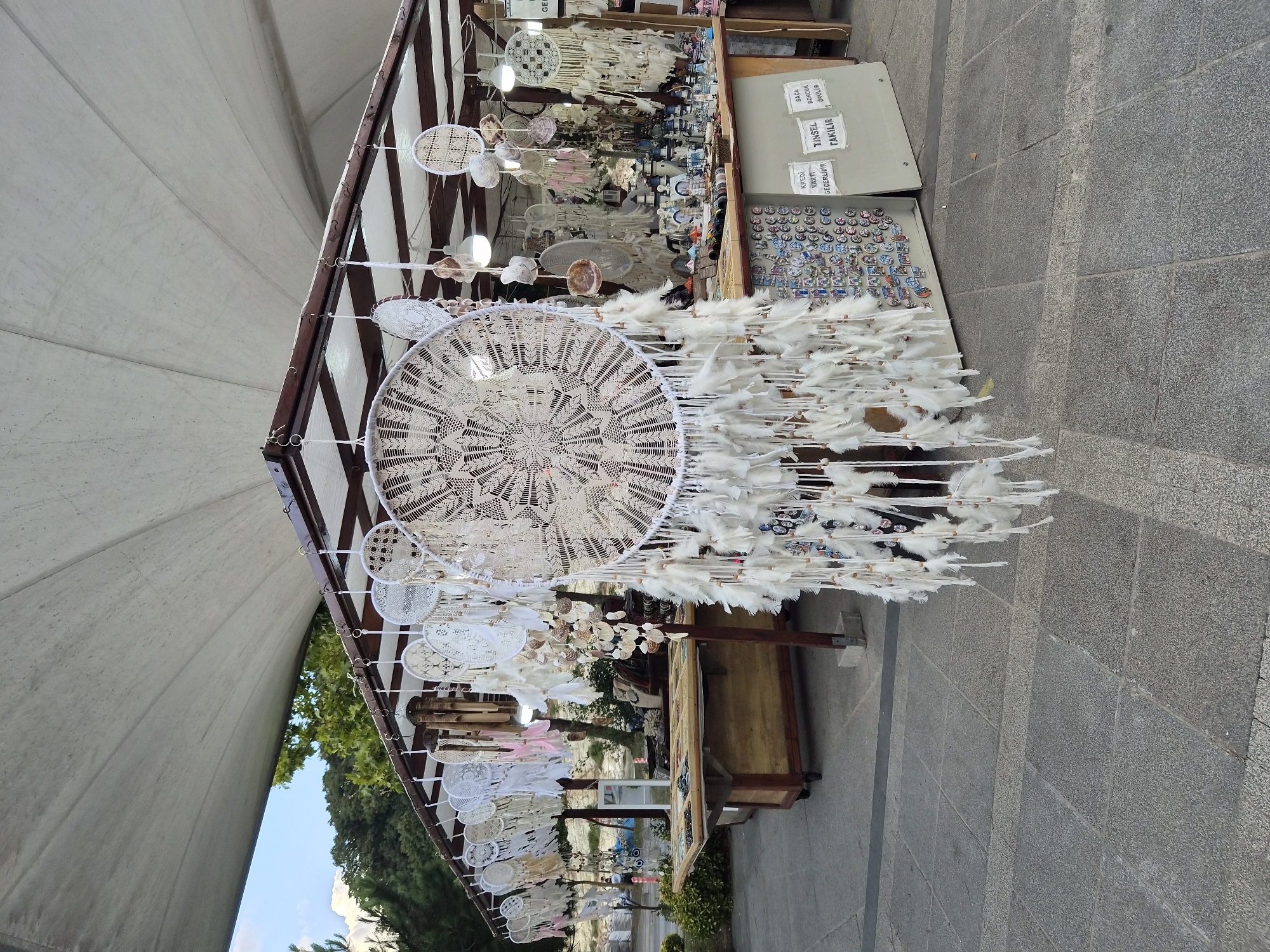The image depicts an outdoor stall in Şile, Turkey, likely a tourist market, showcasing a variety of handmade dreamcatchers. The stall is covered by a large, light-colored canopy stretched over a wooden frame. Numerous dreamcatchers, predominantly white and cream-colored, hang from the frame, varying in size and intricate lacework patterns. Some smaller dreamcatchers are adorned with feathers and shells.
In the foreground, a large, prominent dreamcatcher with a complex, circular woven design and numerous white feathers dominates the center of the frame. Behind it, two wooden tables are laden with goods. The table on the left displays an assortment of jewelry and trinkets. The table on the right, partially obscured, appears to hold more decorative items and possibly souvenirs. A portion of a white board with Turkish text is visible on the right side of the stall.
The ground in front of the stall is paved with grey stone tiles. In the background, trees and a hint of greenery suggest the stall is situated in an area with some natural surroundings. The sky is visible above the canopy, showing a mixture of blue and clouds. No people are clearly visible within the frame, but the setup indicates a commercial environment. The overall impression is one of a vibrant, craft-oriented market stall.