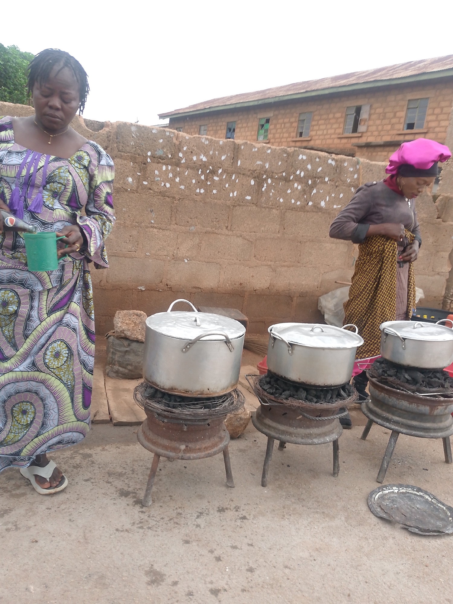 In Bukuru, Nigeria, two women are preparing food outdoors. One woman, wearing a colorful patterned dress, stands to the left, holding a green cup. The other woman, wearing a pink head covering and a brown top with a patterned skirt, stands near the right side of the frame. Three large metal pots sit atop makeshift stoves made from old car rims filled with burning charcoal. The stoves are arranged in a row on a paved surface. Behind them is a low, light-brown brick wall, and a two-story building with a corrugated metal roof is visible in the background.