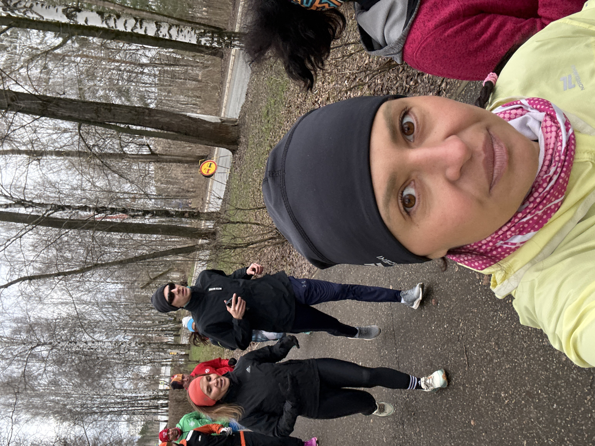 A group of people are on a narrow road, seemingly during a walk or run. The photograph is taken at eye-level by one of the participants. The location is an outdoor environment with tall trees lining the sides of the road. Based on the available clues, the location is Chaikovsky, Russia. Some of the individuals appear to be in athletic wear, like leggings, jackets, and athletic shoes. One person is wearing a black beanie and sunglasses. Another is wearing a red headband. The weather appears to be overcast. The road is paved and there is a forest area on either side. There's a road sign visible, but details are obscured.
