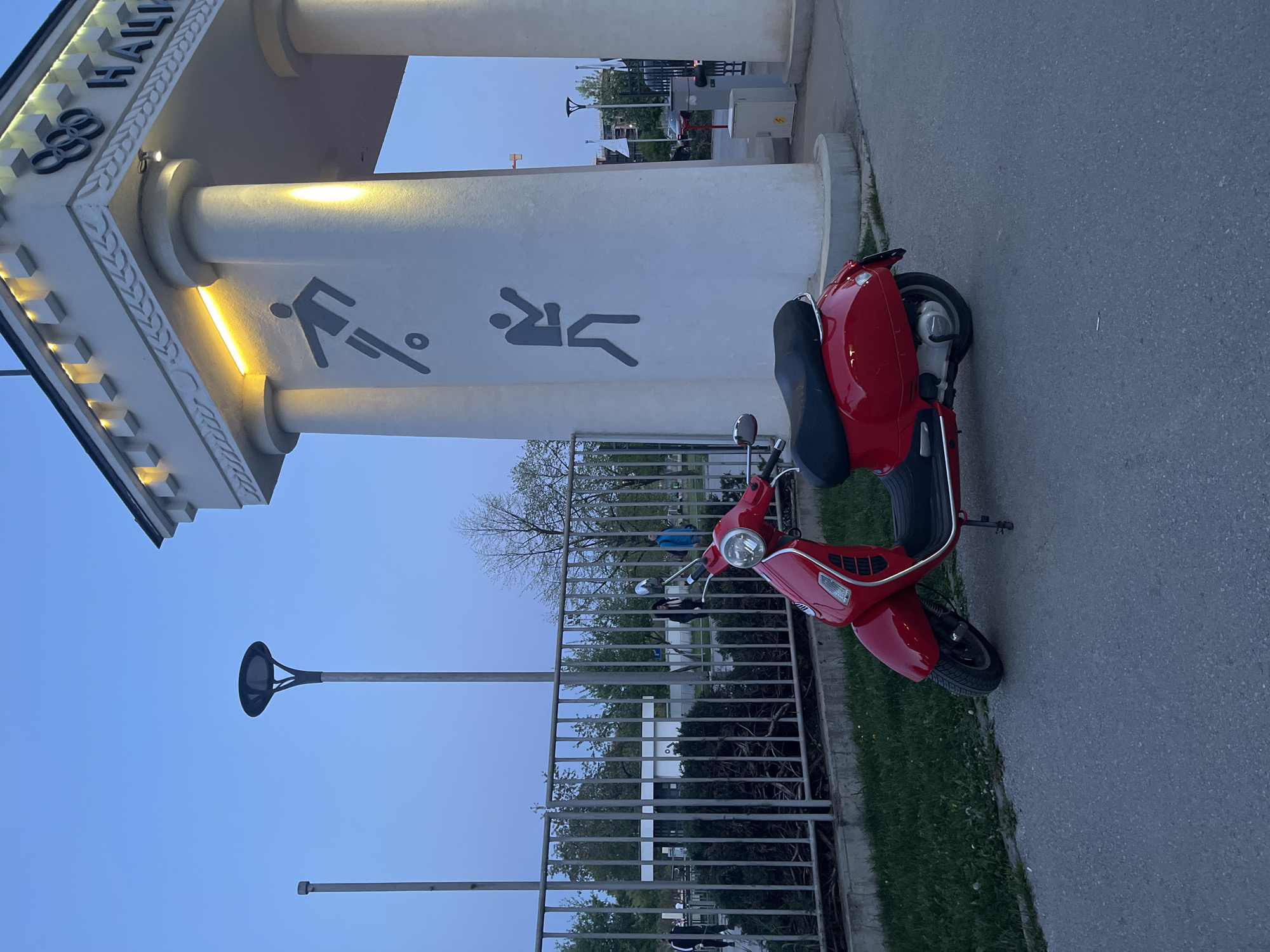 The image captures an outdoor scene at dusk or dawn, likely in Sofia, Bulgaria, characterized by an architectural structure associated with sports and a parked red scooter.
In the foreground, a bright red scooter with a black seat and chrome accents is parked on a grey asphalt surface. To its left, a grey metal fence with vertical bars separates the paved area from a strip of green grass and trees.
Dominating the left side of the frame is a grand, white architectural structure, possibly an entrance archway or part of a building. It features sturdy columns and an ornate frieze. The Olympic rings logo is prominently displayed on the frieze, along with black Cyrillic text that reads "ОЛИ НАЦИ" (likely part of "ОЛИМПИЙСКИ НАЦИОНАЛЕН," meaning "Olympic National"). Warm yellow lights illuminate the underside of the archway and parts of the columns from below. On the side of one column, two dark grey pictograms depicting sports are visible: one of a person kicking a football/soccer ball, and another partially seen figure in an athletic pose.
The sky is a clear, light blue, suggesting late evening or early morning, with artificial lights already activated. Trees are visible beyond the fence, and a modern street lamp with a disc-shaped light fixture stands tall in the distance. The overall atmosphere is calm and peaceful, with no people explicitly visible. The presence of Olympic symbols and sports pictograms strongly indicates the location is related to a national sports complex or Olympic facility.