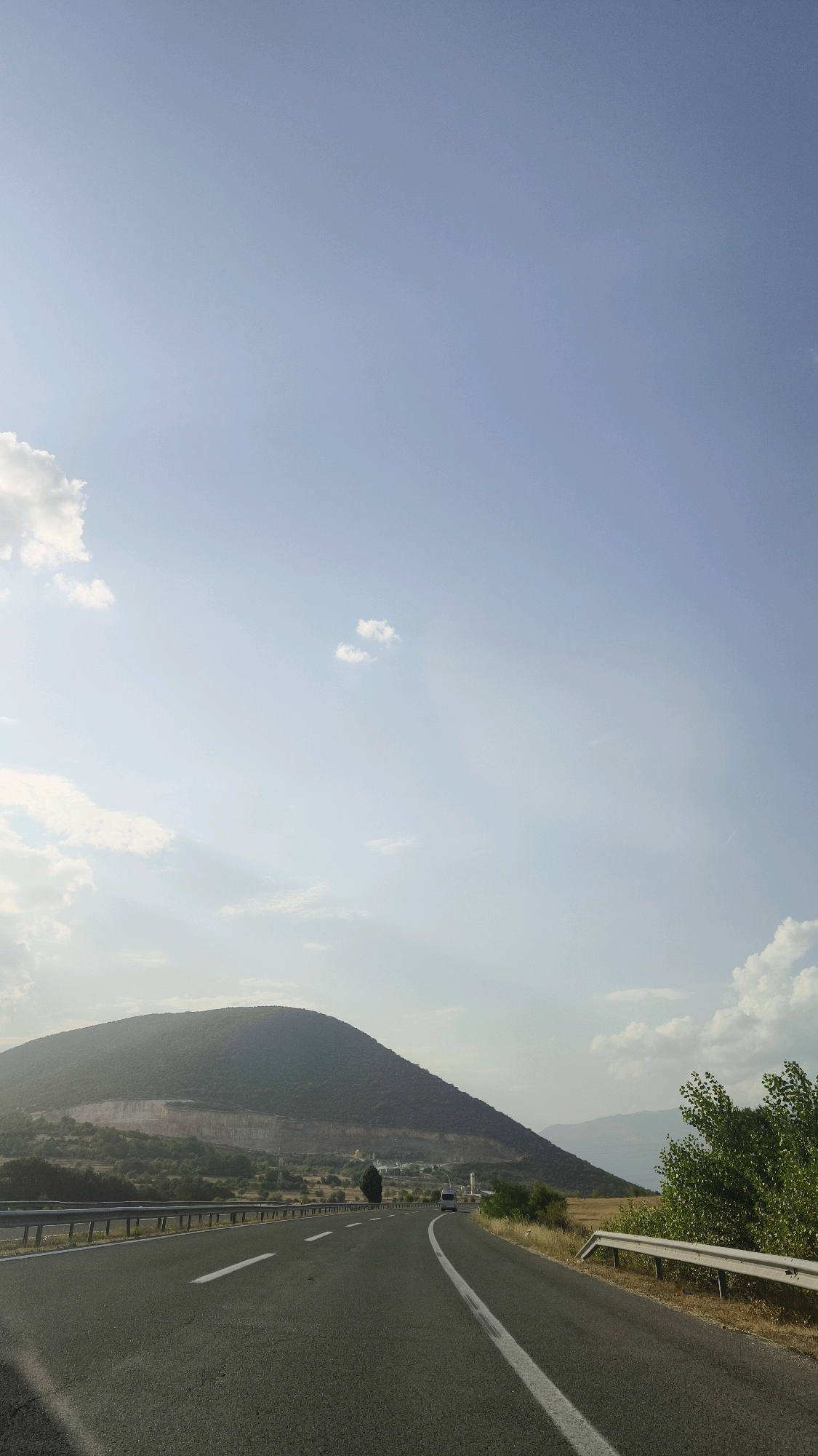 A road stretches into the distance towards a large, tree-covered mountain under a pale blue sky with scattered clouds. The road is paved and has white lane markings. On the left side of the road, a metal guardrail runs alongside the asphalt. The mountain is a dominant feature, its slopes covered in vegetation. In the mid-ground, there is a quarry or cut into the side of the mountain. A small, white van is visible driving on the road in the distance. The environment appears to be rural, possibly near a highway. The time of day appears to be late afternoon or early evening, given the soft lighting. The location is identified as Grupchin, North Macedonia.