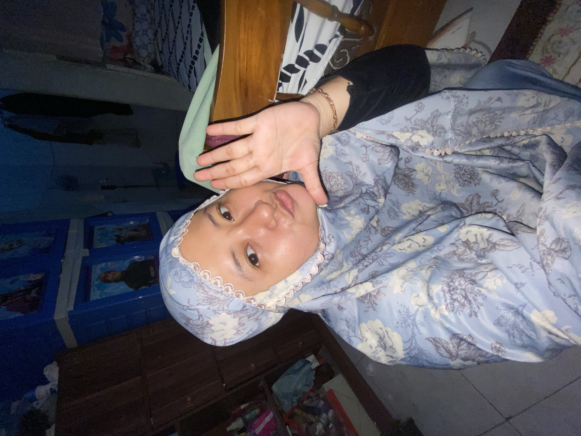 A young woman wearing a blue floral hijab and a black top poses for a selfie in a room. She is holding her right hand up, palm facing the camera, with her fingers spread. Her lips are slightly pursed. Behind her, to the left, is a wooden closet with shelves containing various items. To the right, a bed with a patterned duvet is visible, along with a wooden headboard. The room appears to be dimly lit. The location is stated to be Gelam, Indonesia, though specific details of the environment, such as urban or rural, are not discernible from the image.