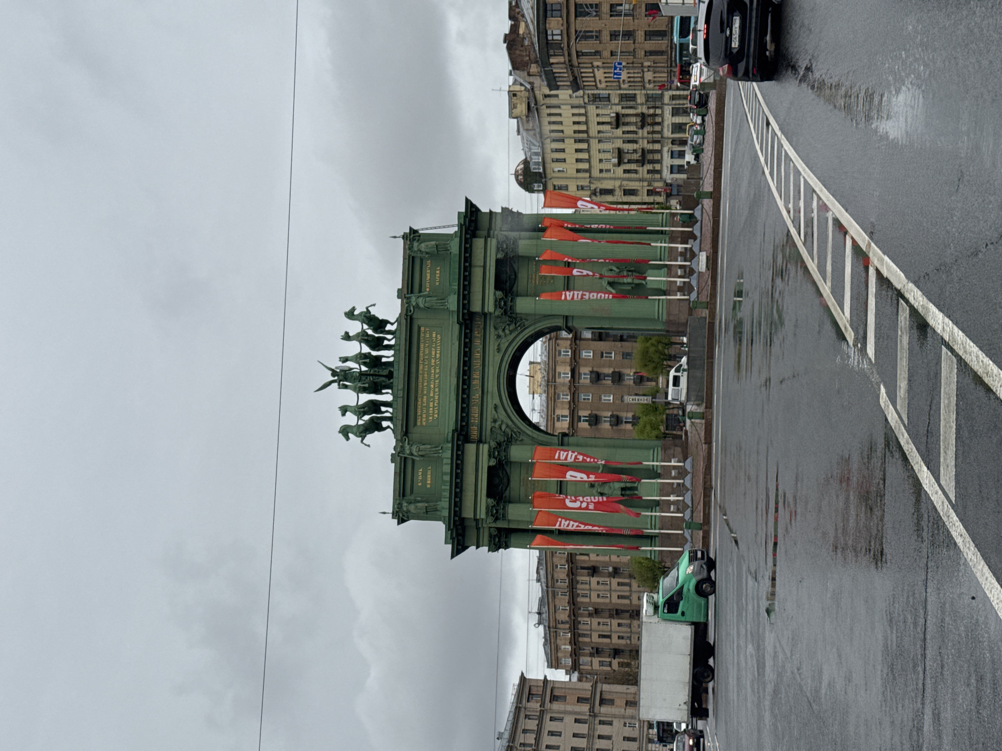 The image captures the Narva Triumphal Arch in Saint Petersburg, Russia, under a cloudy sky. The arch, constructed of green metal, features a quadriga atop its structure. Red flags, adorned with white text, are positioned along the base of the monument. Behind the arch, buildings stand tall. Wet asphalt covers the street, reflecting the surrounding environment. A green delivery truck is parked nearby, and vehicles travel on the road. The presence of lane markings indicates traffic lanes. No people are readily visible in the immediate foreground.