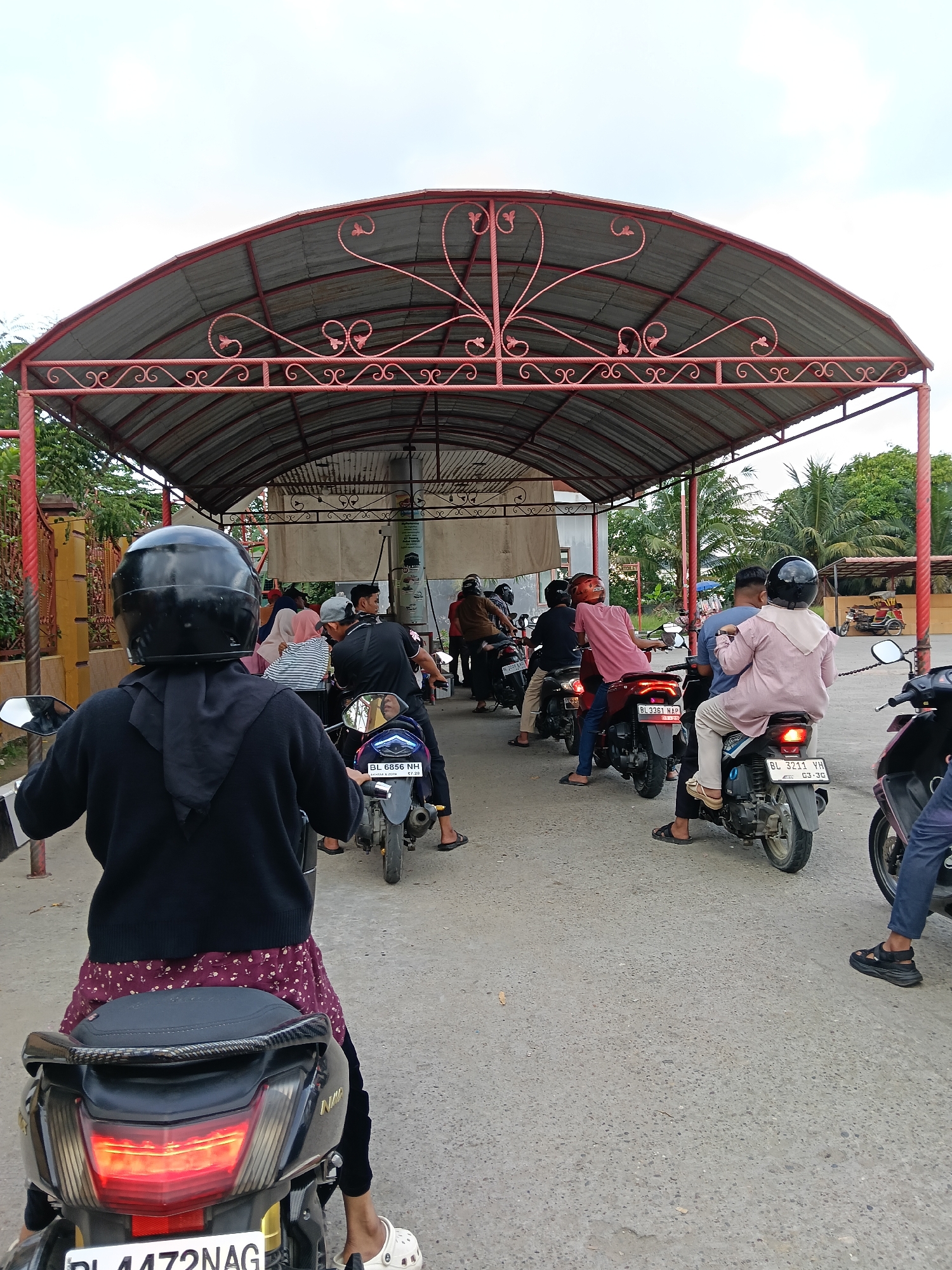 The image depicts a line of people on motorcycles waiting under a red metal awning. The location is likely Lhokseumawe, Indonesia, as suggested by the license plates on the motorcycles. The motorcycles are of various makes and models. The individuals are of different ages and genders. The environment appears to be a street or parking area.