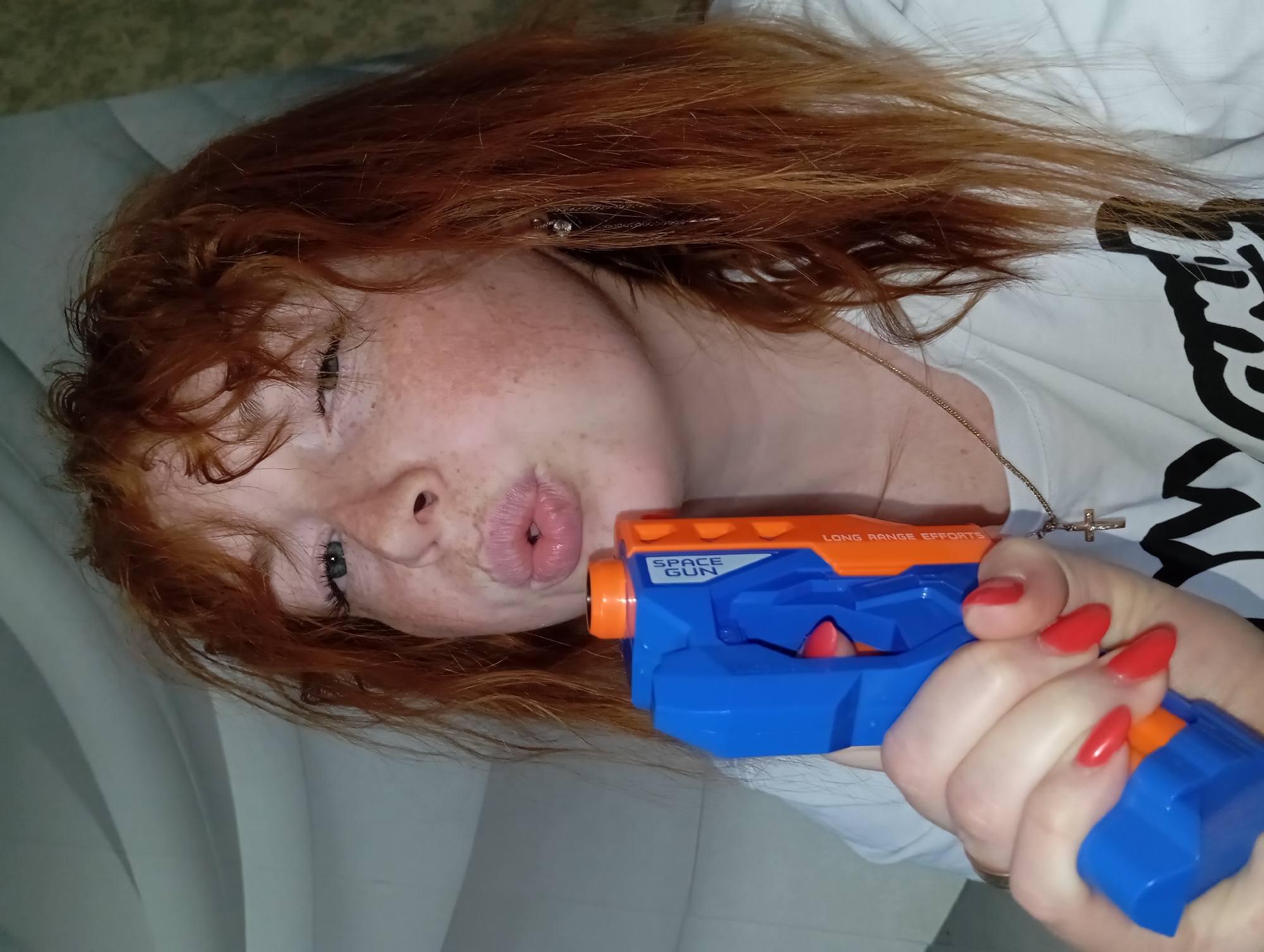 A person with red hair and freckles is lying on a bed, holding a blue and orange toy gun labeled "SPACE GUN" in front of their face. Their lips are pursed. The person is wearing a white shirt with black designs. A silver necklace with a cross pendant is visible. The background is a light-colored sheet. The location is Unknown, Unknown.