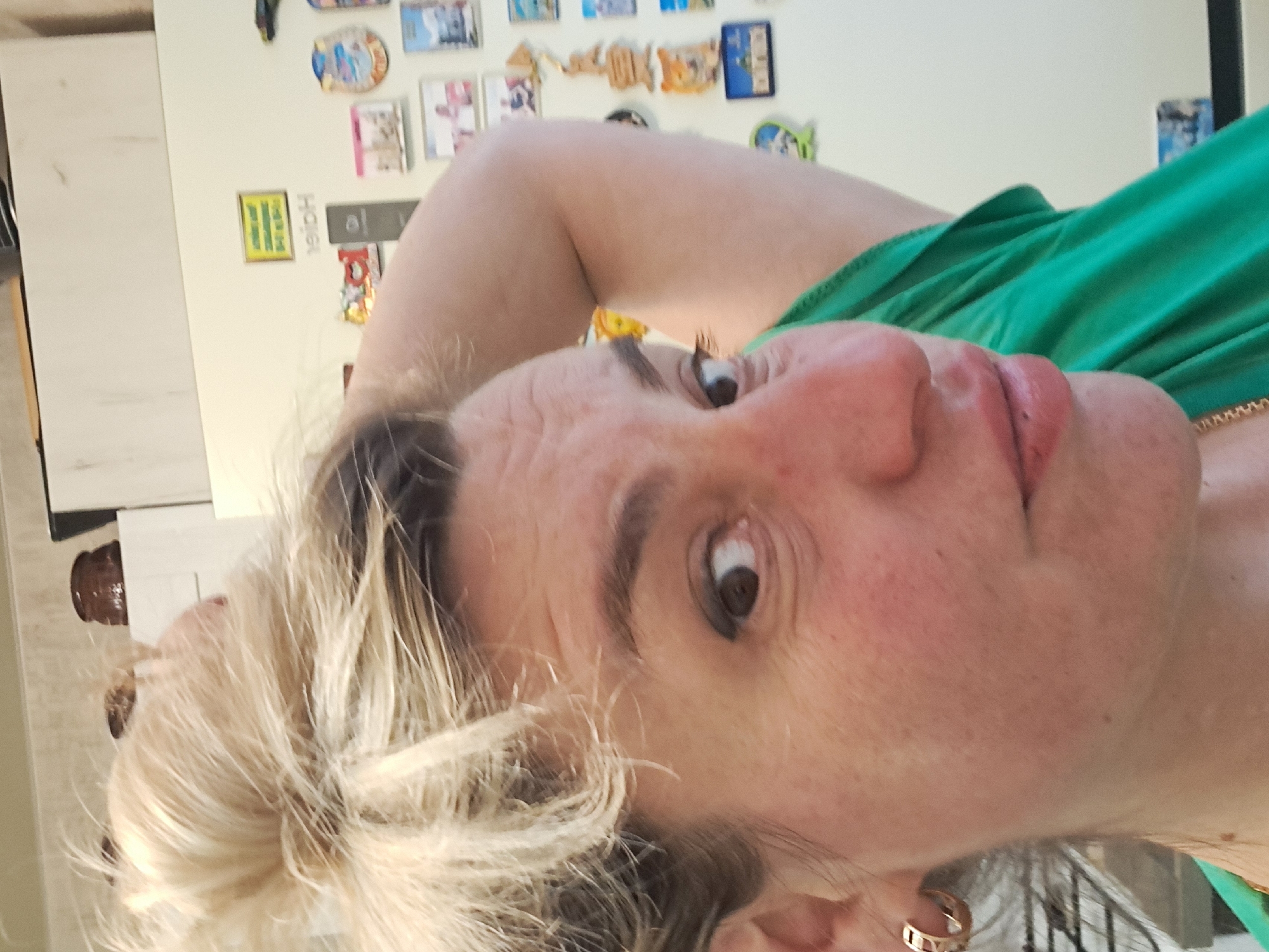A selfie of a middle-aged woman with blonde hair and a green top. She is positioned close to the camera, her head tilted. Behind her is a refrigerator covered with various magnets and stickers. The location in Elektrostal, Russia is not determinable from the image.