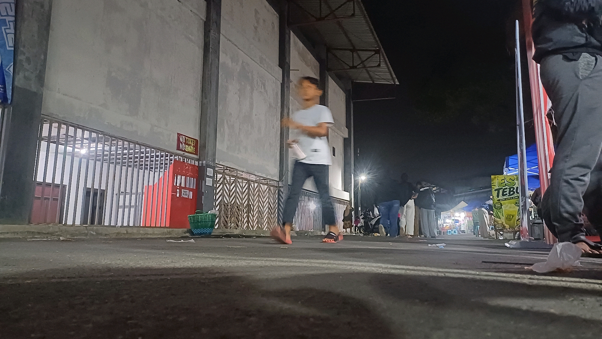 The image depicts a night scene in the city of Kowangan, Indonesia. The foreground is dominated by a dark asphalt ground, with some scattered white debris. In the mid-ground, a young male in a light-colored t-shirt and dark pants is walking from left to right, appearing to be in motion. To the left, a red structure with metal bars in front of it is visible, featuring signs that read "NO TIKET NO GAME" and "TIKET Rp. 20.000". A green woven basket sits near the red structure. Further to the right, in the background, a lively street scene unfolds with tents, stalls, and a crowd of people. A prominent sign with the text "ES TEBU" and an image of a beverage is also visible in the background. The overall environment suggests a public gathering or event at night.