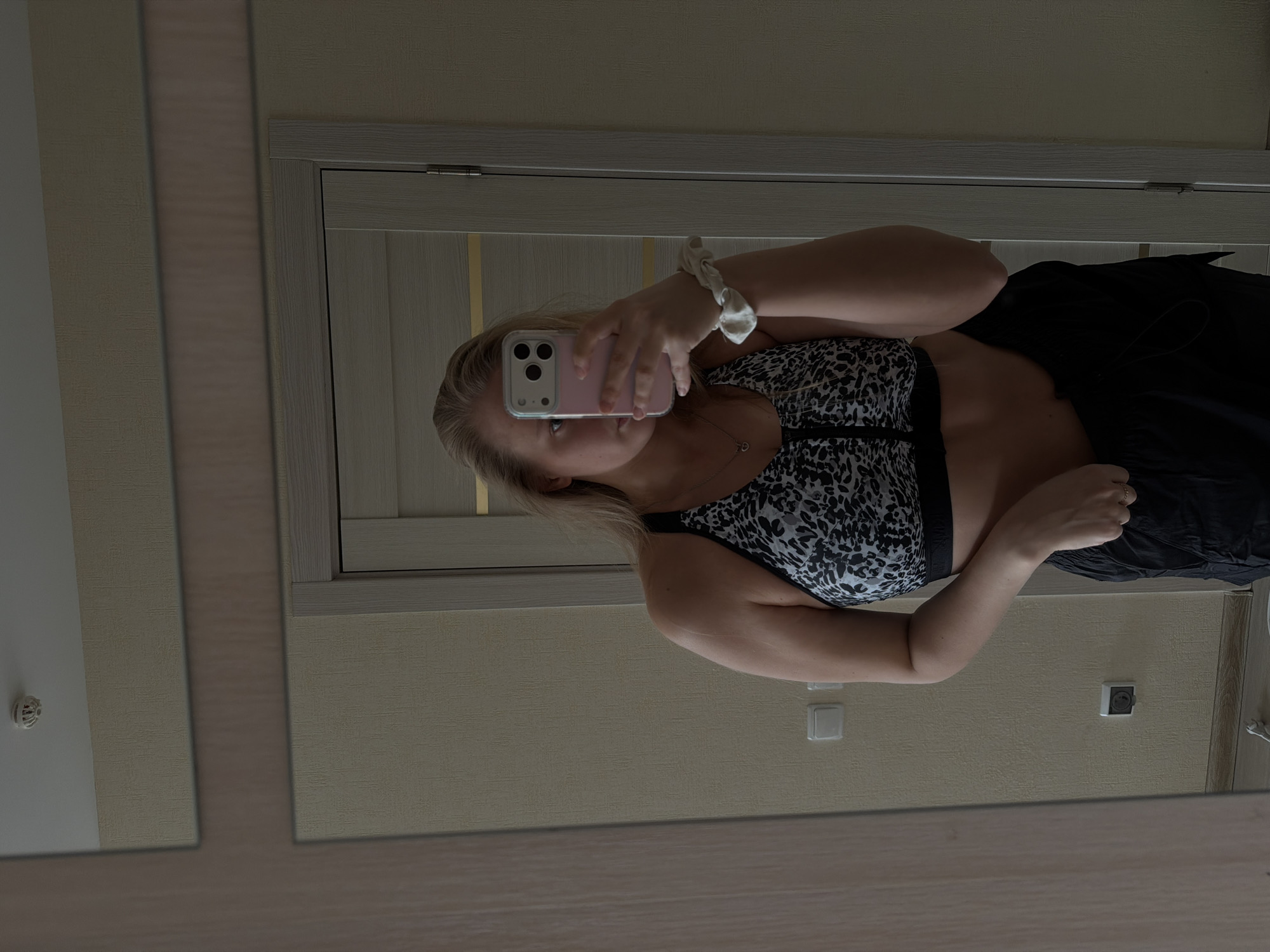 A woman is taking a selfie in front of a mirror. She is wearing a patterned sports bra and black bottoms. She is holding a pink smartphone up to her face. Her hair is blonde and pulled back with a white scrunchie. The background appears to be a room with light beige walls and a white door frame. There is an electrical outlet visible on the wall behind her. There is no indication of the location being in Izhevsk, Russia.