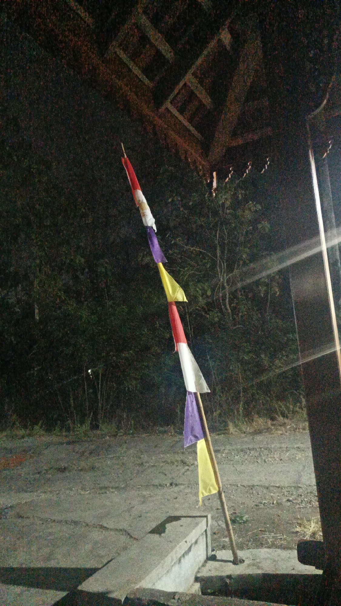 The image depicts a wooden pole adorned with a string of colorful triangular flags. The flags are arranged sequentially in alternating colors of red and white, followed by purple and yellow. They are tied to the pole at varying intervals, creating a festive or decorative display. The pole is set against a backdrop of dark foliage, suggesting an outdoor environment, possibly in a rural or semi-urban setting. In the foreground, a concrete structure, possibly a pathway or a low wall, is visible, casting a shadow. The overall lighting suggests it is nighttime or a dimly lit area. The location is identified as Sambi, Indonesia.