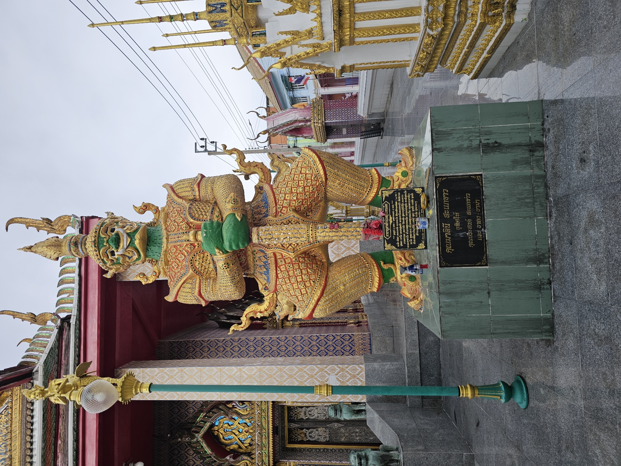 A towering, ornate golden statue of a green-faced guardian figure, likely a Yaksha, stands prominently in front of a building with a red roof and intricate golden decorations. The statue is depicted in a seated position, holding a green object and a sword. A plaque with Thai script is affixed to the base of the statue. The scene is set outdoors, on a paved surface, with other architectural elements and some electrical wires visible in the background. The statue appears to be a significant cultural or religious monument, possibly located within a temple complex.