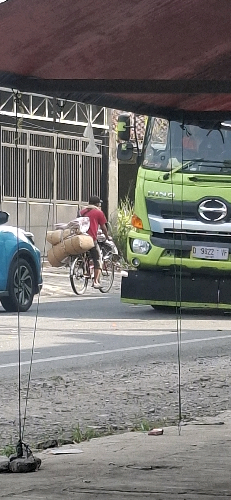 A person is riding a bicycle laden with a large bundle of goods. The bicycle is on a paved road in front of a modern building with vertical slats and a white roof structure. To the right of the bicycle is a bright green HINO truck. The scene appears to be in the city of Rancagoong, Indonesia. A blue car is partially visible on the left side of the image. The background shows a street with a rough textured surface. The image is framed by a dark red awning at the top and thin vertical wires in the foreground.