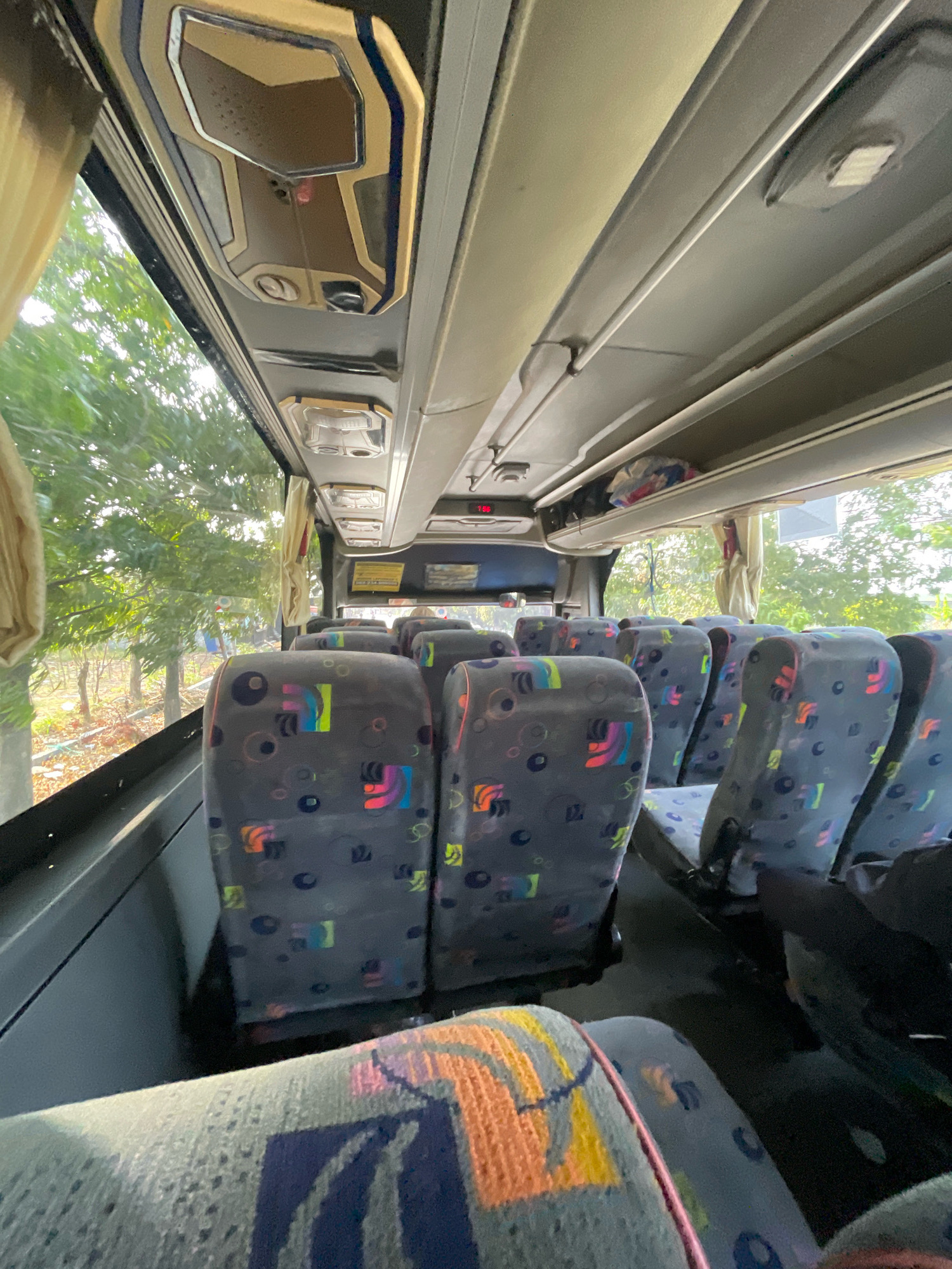 The image depicts the interior of a passenger bus, likely in Sampang, Indonesia. The perspective is from a seat looking forward and upward. The seats are upholstered in a gray fabric with a colorful abstract pattern of circles and geometric shapes. Overhead compartments are visible, with luggage stowed in one. The ceiling features interior lighting fixtures and ventilation. Sunlight streams in from the windows on the left side, illuminating trees and foliage outside. The floor of the bus is dark. The overall impression is a standard interior of a public transportation vehicle.