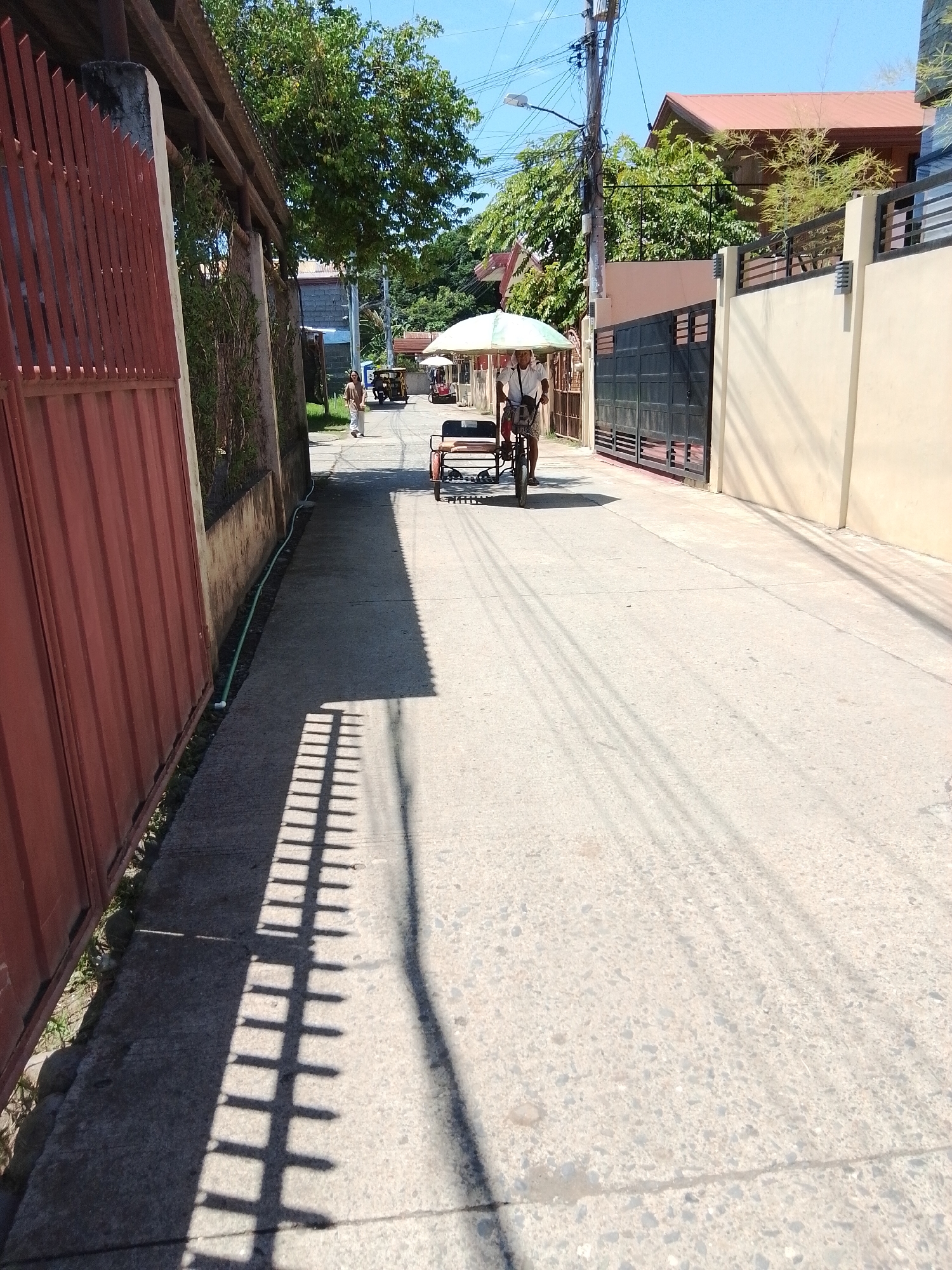 The image depicts a sunny day in a residential area of Davao City, Philippines. A narrow asphalt road runs through the scene, flanked by residential houses and fences. The perspective is from a ground-level viewpoint, looking down the street.
On the left side of the road, a tall, red corrugated metal fence lines the property. Further down the street, a person is walking away from the camera, wearing light-colored clothing.
In the middle of the road, a man is riding a tricycle with an umbrella attached for shade. The tricycle appears to be a common mode of transport in the Philippines, often adapted for passenger or cargo use. The man is wearing a white shirt and patterned shorts.
The environment is typical of a tropical urban setting, with lush green trees visible in the background and clear blue skies overhead. Electrical wires are strung across utility poles, a common sight in Philippine cities. The sun casts distinct shadows on the road and sidewalk, creating a pattern of light and shade that adds depth to the image. The houses have distinct architectural styles, with visible roofs and walls. One house on the right features a prominent beige-colored wall and a dark metal gate. The overall impression is one of a peaceful, ordinary day in a Philippine neighborhood.