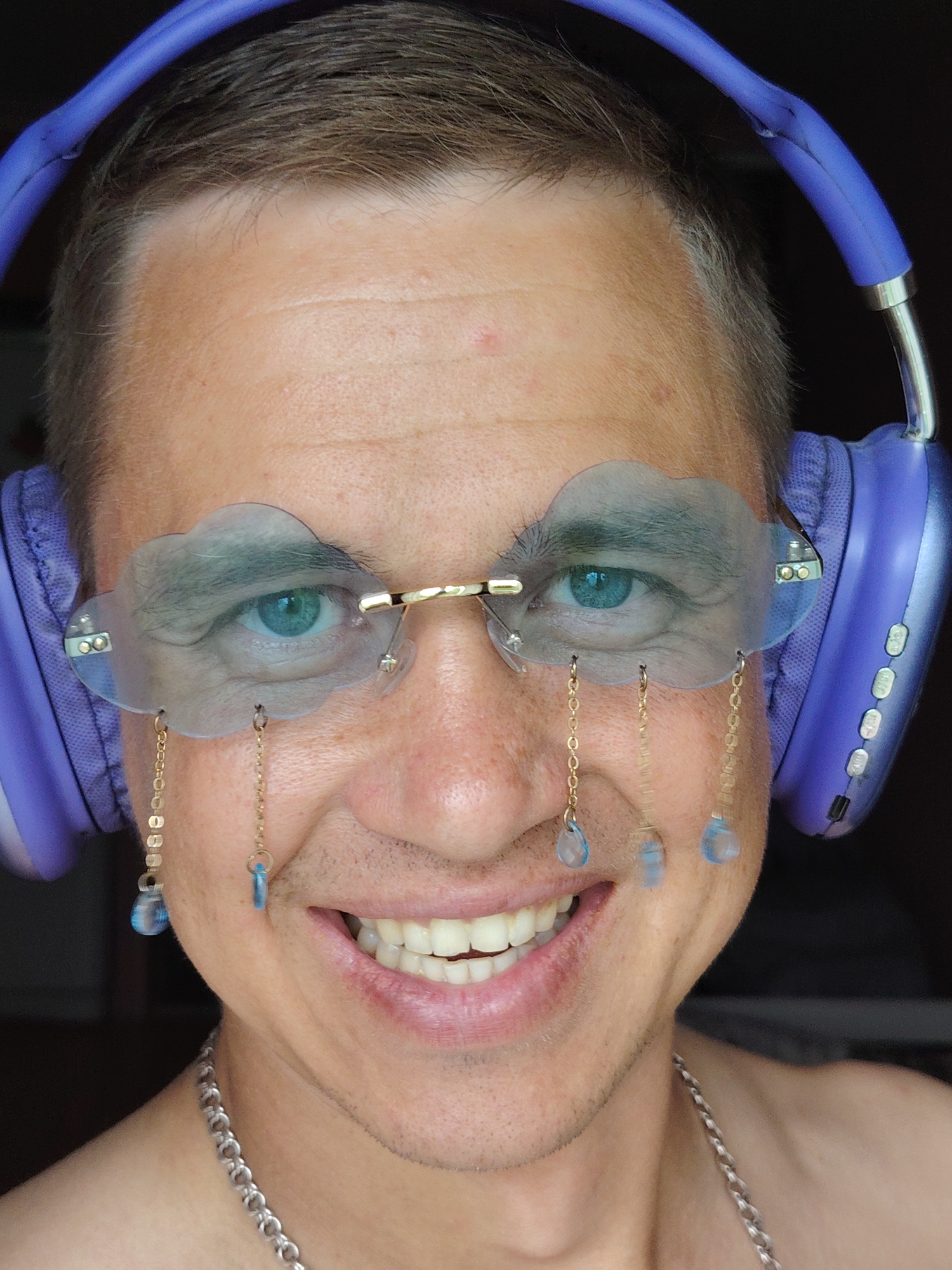 An adult male is depicted in a close-up, head-and-shoulders portrait, facing the camera with a direct gaze and a wide, open-mouthed smile revealing white teeth. He has light skin, short brown hair with hints of grey, and visible stubble on his lower face. He wears large, over-ear headphones of a light purple color, which show a tear on the headband above his right temple. His eyes are bright blue. He is wearing novelty rimless glasses with lenses shaped like light blue clouds. From the bottom edge of each cloud lens, multiple gold-colored chains hang, each terminating in a small, translucent blue teardrop bead, simulating rain. A silver-colored chain necklace is visible around his neck. The background is dark and out of focus, providing no discernible environmental details. The location in the city of Chaikovsky, Russia, is not identifiable from the visual content of the image.