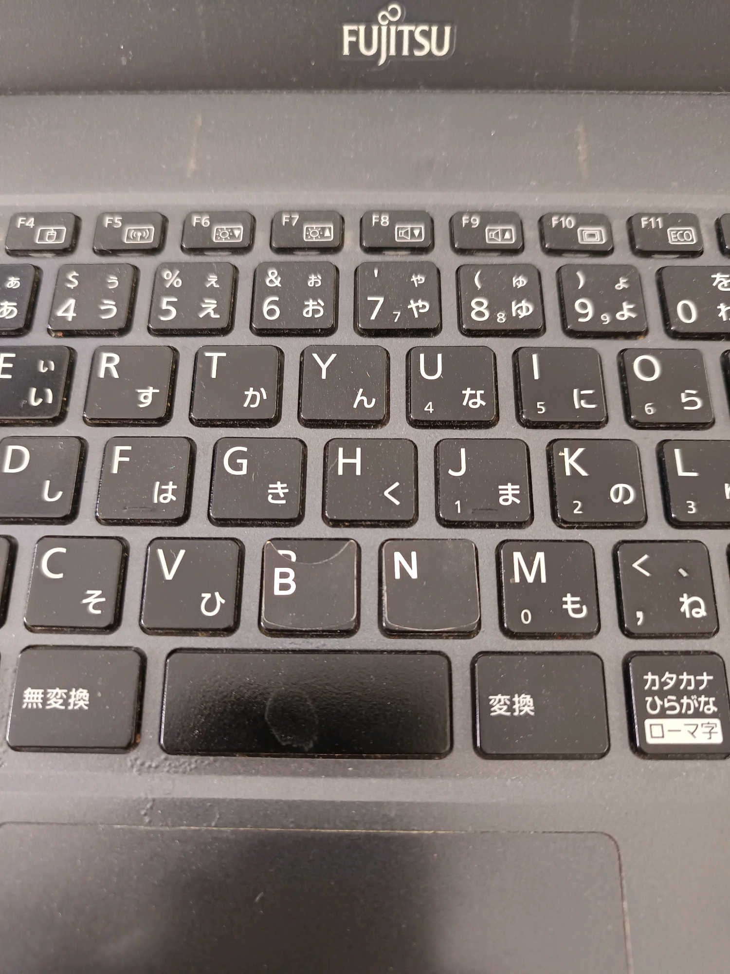 The image shows a close-up, overhead view of a dark gray laptop keyboard. The keyboard is made of black plastic keys with white lettering. The brand name "FUJITSU" is visible at the top of the keyboard. The keys display a mix of English letters and Japanese characters, indicating it is likely a Japanese keyboard layout. Some keys have multiple characters or symbols on them, with smaller characters above the main ones, suggesting a shift or modifier function. The keys are arranged in a standard QWERTY layout for the English letters, but the Japanese characters are superimposed. Specifically, the row below the function keys (F1-F12) contains Japanese characters corresponding to numbers and symbols. The rows below that contain English letters alongside Japanese characters. For example, the 'A' key has 'a' and 'い' (i), the 'S' key has 's' and 'う' (u), and the 'D' key has 'd' and 'え' (e). Further down, the 'J' key has 'j' and 'ま' (ma), and the 'K' key has 'k' and 'の' (no). Special function keys are visible at the bottom, including "無変換" (mu henkan - conversion), "変換" (henkan - conversion), and "カタカナ ひらがな ローマ字" (katakana hiragana romaji - Katakana, Hiragana, Romaji). The keyboard appears slightly worn, with some scuff marks and a smudged area on the spacebar. There are no people or other objects clearly visible in the image beyond the keyboard itself. The background is out of focus and appears to be the dark surface of the laptop casing. There is no information in the image to suggest a location in Labuhan, Indonesia.