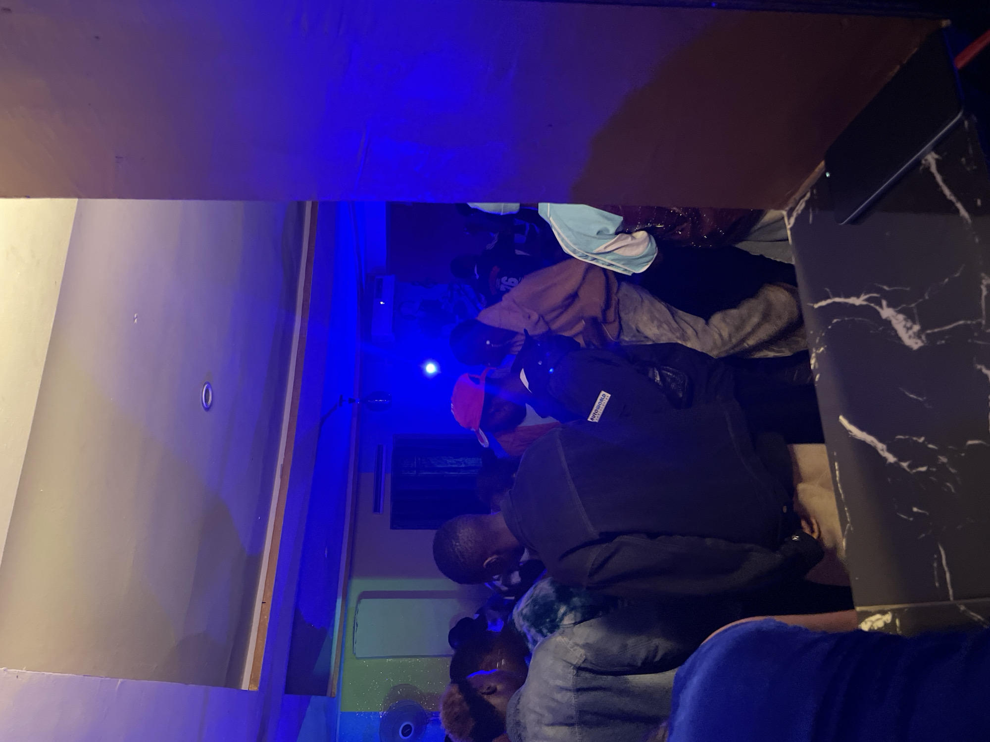 In Durumi, Nigeria, a dimly lit indoor gathering takes place, with a strong blue ambient light casting a vibrant hue across the scene. A group of individuals are visible, some standing close together, suggesting a social interaction or event. Clothing appears casual, with jackets and shirts being the primary visible attire. The environment includes a ceiling with visible lighting fixtures and a wall that partially obscures the view, creating a sense of depth. In the foreground, a counter with a black and white marbled surface is present, and a dark smartphone rests on it, its screen illuminated by a faint light. The overall atmosphere suggests a lively social setting, possibly a club or party, characterized by the interplay of light and shadow and the presence of a crowd.