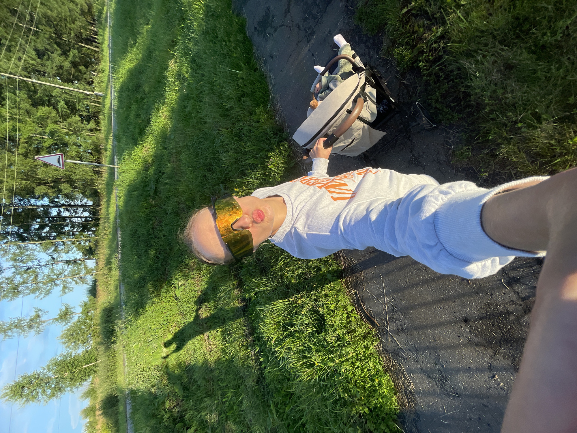 This image depicts a person pushing a stroller on a paved path next to a grassy area. The person is wearing a white sweatshirt, sunglasses with reflective, amber lenses, and is looking towards the camera. The stroller, which appears to contain a child, is positioned in front of the person. The setting is outdoors, with lush green grass and trees visible in the background. A road sign is also present in the distance. The lighting suggests it is daytime, likely late afternoon due to the long shadows cast. The overall mood appears casual and relaxed, possibly indicating a leisurely walk. The image is oriented sideways.