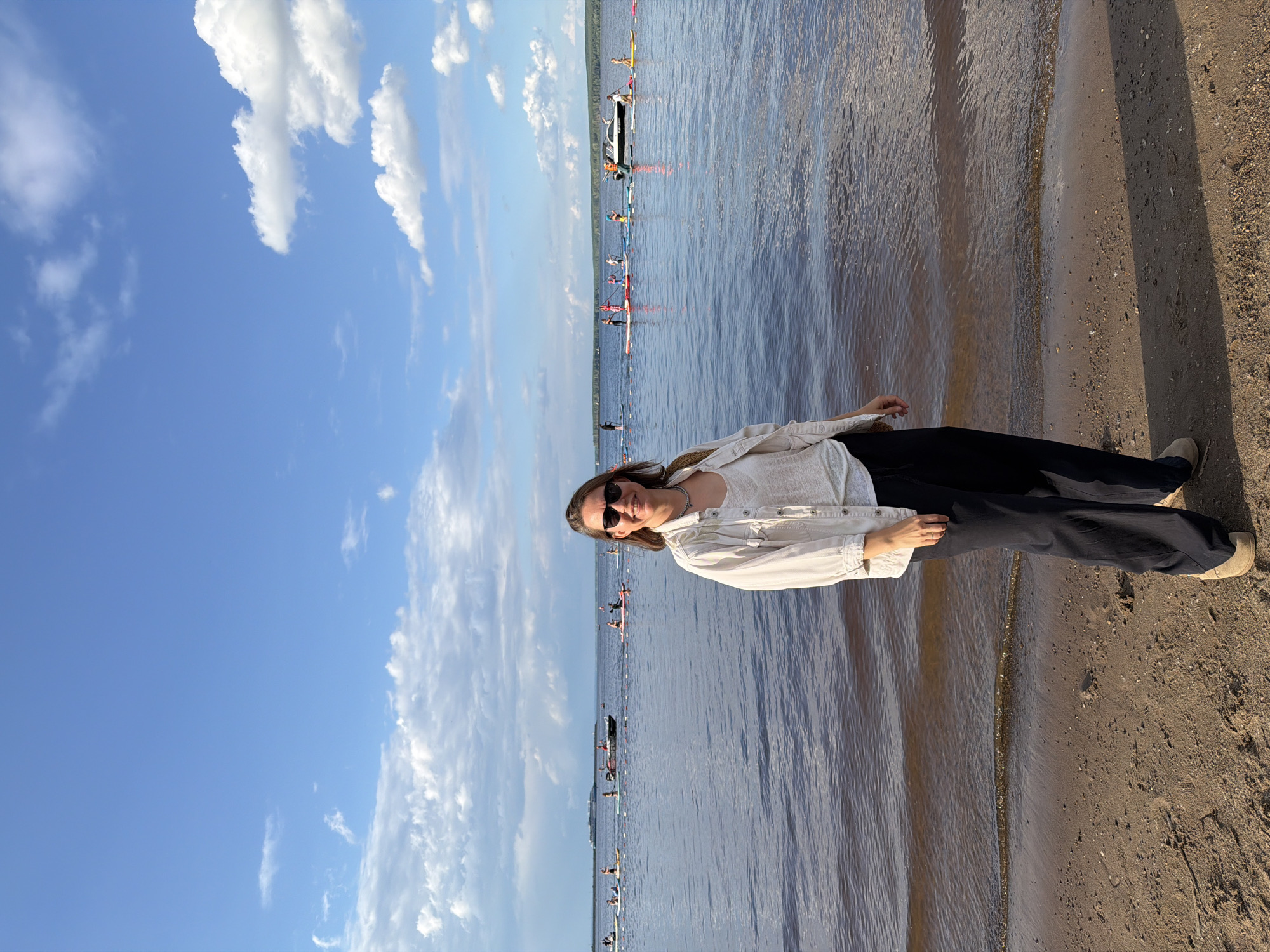 A full-body, eye-level shot captures a woman standing on a sandy shore, facing the viewer. She is wearing sunglasses, a light-colored unbuttoned shirt over a white top, and dark, loose-fitting pants. Her right hand rests on her hip, and her left arm is down. Behind her, a calm body of water stretches to the horizon, with a faint line of trees visible in the distance. Several people on paddleboards are scattered across the water, some with paddles raised. A white motorboat with people on board is also present on the water. The sky is a bright blue, dotted with white, puffy clouds. The scene appears to be located on the Kama River in Chaikovsky, Russia, possibly during a recreational event or competition.
