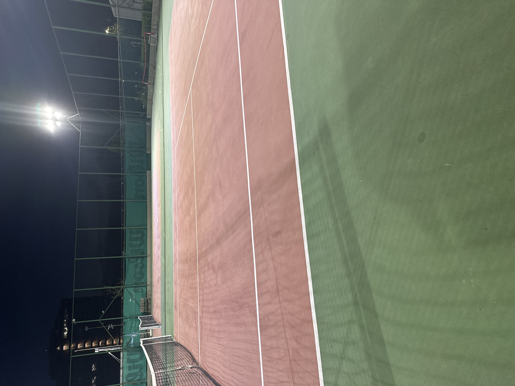 An outdoor tennis court at night, illuminated by overhead lights. The court surface is a reddish-brown color with green out-of-bounds areas, marked by white lines. A tennis net divides the court in the foreground. In the background, a green fence surrounds the court, with a building visible beyond it. The image is taken from a low angle, looking across the court. The location is not specified in the image.