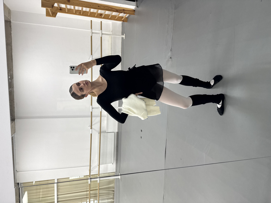 A female subject is captured in a mirror selfie within a dance studio, inferentially located in Tbilisi, Georgia. The subject, an adult female with light brown hair secured in a low style, is dressed in a black long-sleeved leotard, a sheer black wrap skirt, white ballet tights, black leg warmers, and black ballet shoes. She holds a folded cream-colored ribbed sweater draped over her left forearm. A light-colored smartphone with a multi-lens rear camera is held in both hands, positioned to capture her reflection. The studio environment features a large full-length mirror occupying the foreground wall. In the background, two parallel wooden ballet barres are mounted on a white wall. A white radiator is visible below a window with sheer curtains on the left side, while a wooden wall-mounted ladder-like gymnastic apparatus is present on the right. The floor is light gray, and the ceiling consists of exposed concrete. The subject stands with her body slightly angled, looking towards the phone.