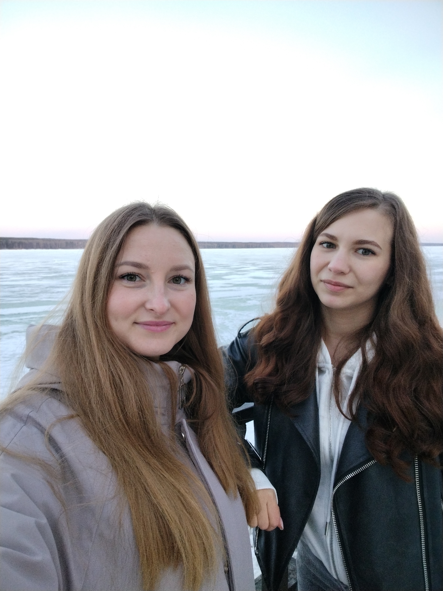 Two young women are posing for a selfie outdoors. The woman on the left has long, light brown hair and is wearing a light gray jacket. The woman on the right has long, dark brown hair and is wearing a black leather jacket over a white hoodie. The background appears to be a frozen lake or river. In the distance, a forest line is visible. The sky is a pale, overcast color. The image was taken in the city of Chaikovsky, Russia.