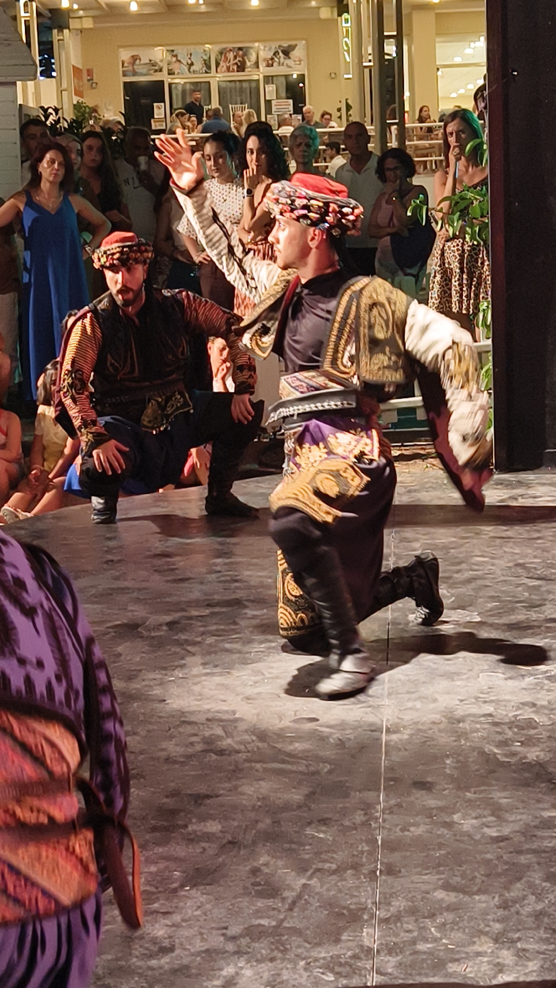 Two men in traditional Turkish clothing are performing a dance on an outdoor stage in Kuşadası, Turkey. The man in the foreground is kneeling on his right knee with his left leg extended forward, and his right arm is raised in the air, palm open. He wears a dark vest over a patterned shirt, a sash around his waist, and dark pants tucked into boots. His head is adorned with a red and multi-colored headdress. To his left, another man is crouched low, facing the viewer. He wears a similar vest over a patterned shirt and dark pants. A crowd of spectators is visible in the background, some sitting and some standing. The stage appears to be made of concrete, and there are some overhead lights and decorations visible. The setting suggests a cultural performance or festival.