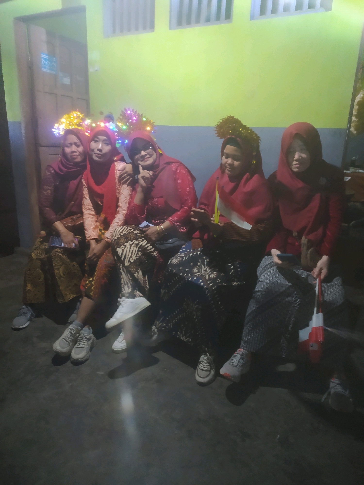 Five women are seated indoors, facing forward. They are all wearing dark red headscarves, and most are also wearing dark red tops and patterned skirts. Several of the women have elaborate headpieces adorned with colorful lights. They are posed together, with some of the women touching each other. In the foreground, their legs and feet are visible, and they are all wearing sneakers. The background consists of a dimly lit room with a lime green wall. The location is specified as Karanggayam, Indonesia. The women appear to be participating in a celebratory event or gathering.