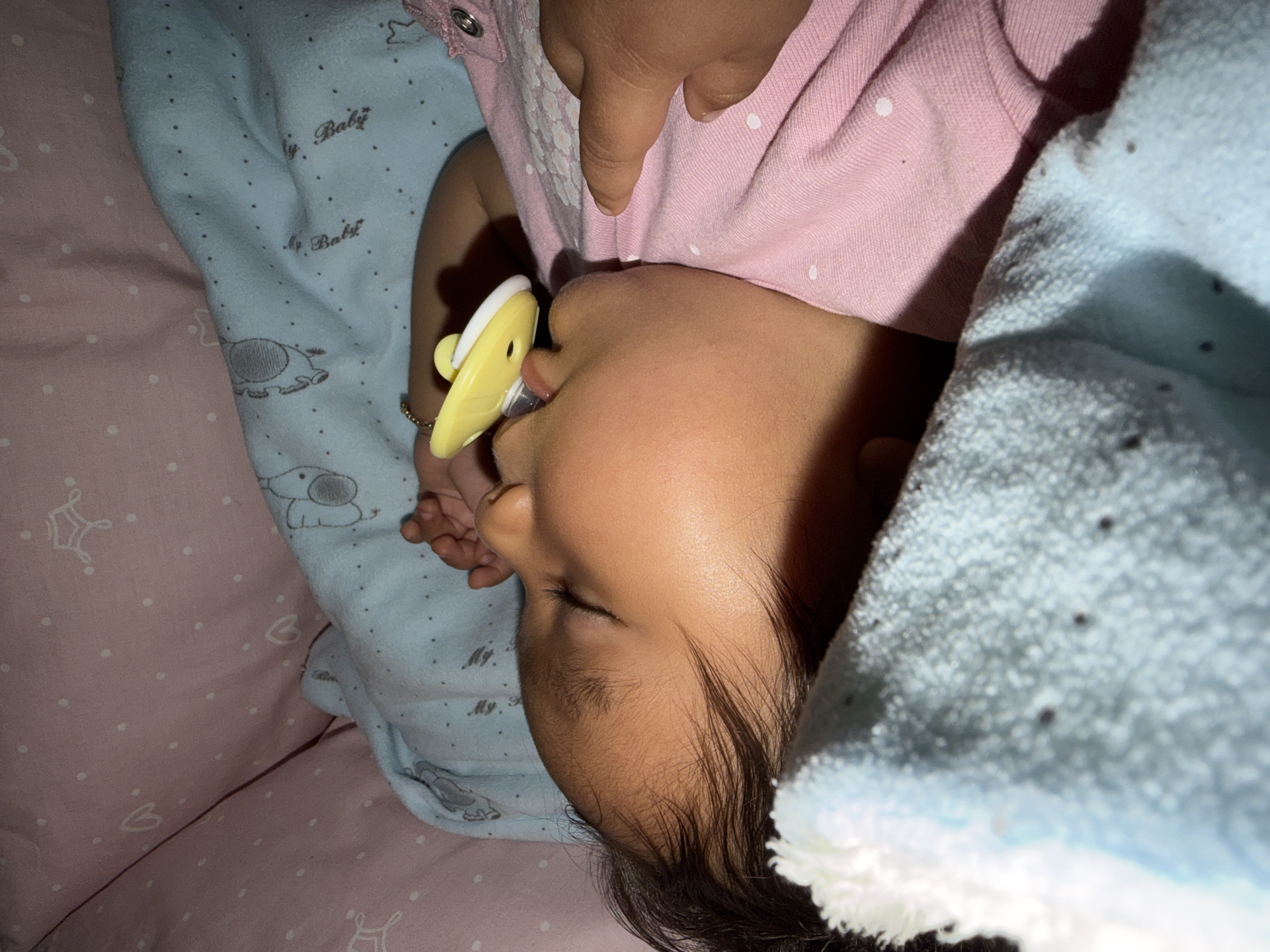 A close-up, overhead shot shows a sleeping baby lying on its side with a pacifier in its mouth. The baby is nestled in a crib with pink and blue patterned blankets. The blankets are adorned with small white dots and images of elephants. A hand is visible on the right side of the frame, near the baby's chest. The baby is wearing a pink onesie. The lighting is dim, suggesting it is nighttime. This scene is within the city limits of Razhevo Konare, Bulgaria.