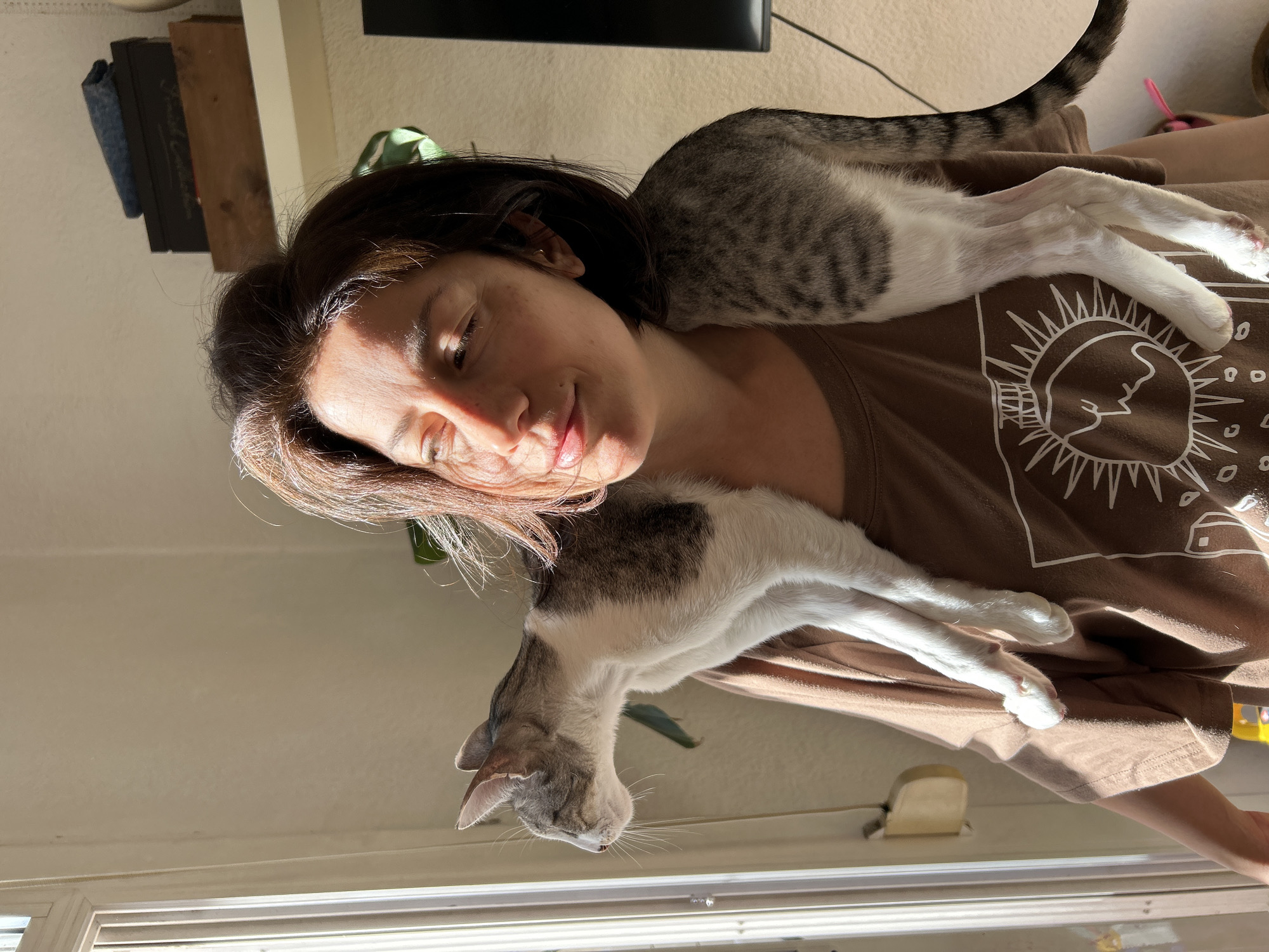 The image portrays a peaceful indoor scene in Barcelona, Spain, featuring a woman resting with two cats. A woman with dark hair lies on her back, her head tilted, eyes gently closed or partially shut, and a soft, content smile on her face. Bright natural sunlight streams in, illuminating her face and the animals.
Two grey and white cats are draped across her body. One tabby cat stretches across her shoulder and upper back, its front paws extending upwards, with its striped tail visible at the top. The second grey and white cat rests on her chest and torso, its head near her left arm. Both cats appear relaxed and comfortable, mirroring the woman's serene state.
The woman is wearing a brown t-shirt featuring a prominent white graphic design of a sun with a stylized face and radiating lines. In the background, a shelf holds various items, including a dark object partially labeled "Vogue Collection," a wooden block, and a blue fabric item. A glimpse of a window or blinds is visible at the bottom of the frame, and a green plant leaf can be seen subtly near the second cat. The strong, warm lighting suggests it is daytime, likely morning or late afternoon, creating an ambiance of calm and domestic companionship.