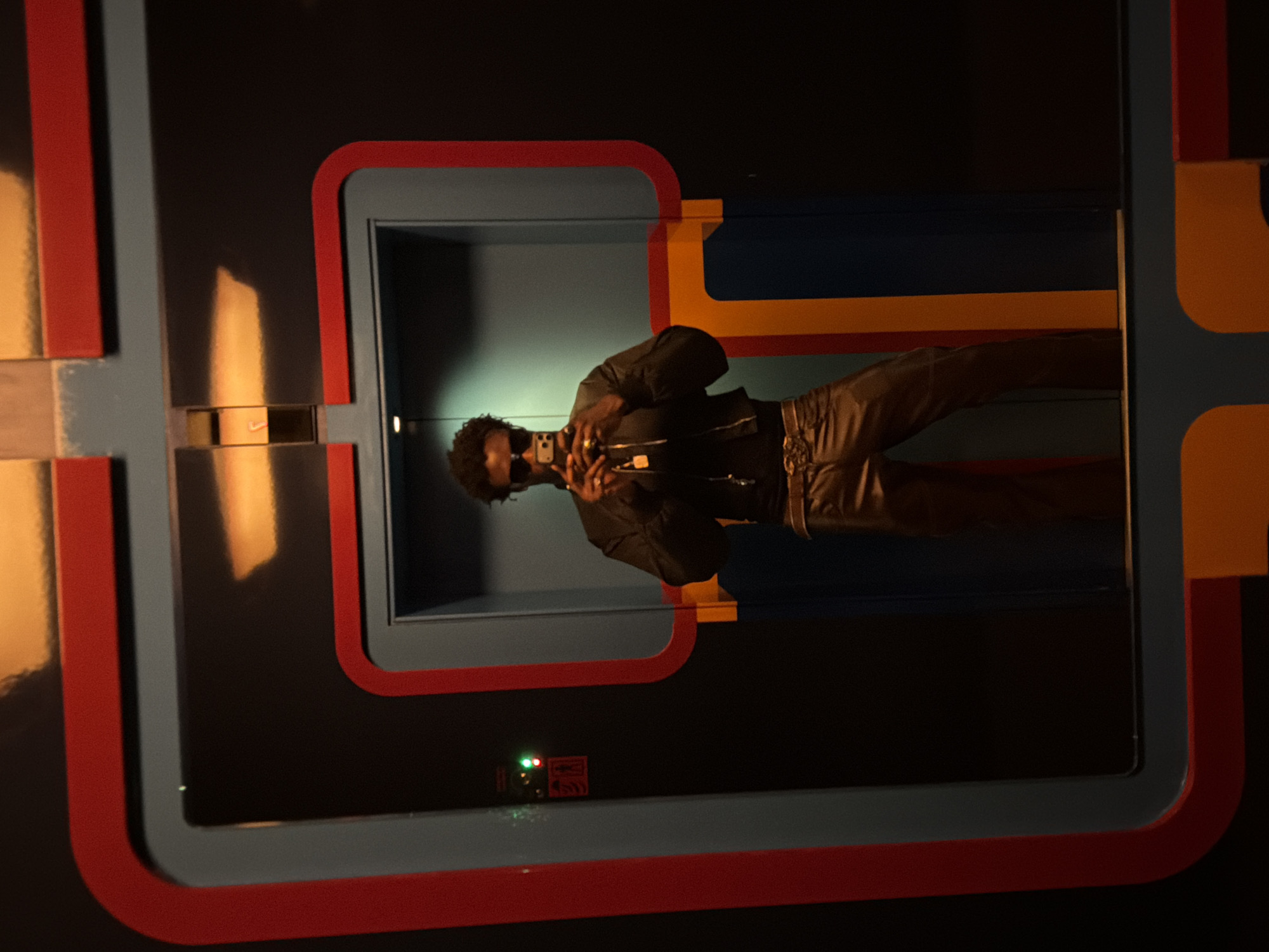 A person in a black jacket and brown pants is taking a picture of themselves in a rectangular, multi-layered frame consisting of red, light blue, and black colors, reminiscent of a retro arcade game aesthetic. The frame contains a light blue section that resembles an elevator or small room. The person is in the room, angled to the side, holding a phone or camera in their hands. There's a small green light and a rectangular object with symbols near the bottom part of the light blue section. The overall environment appears dark, with some reflective surfaces casting light patterns. The location is London, United Kingdom.