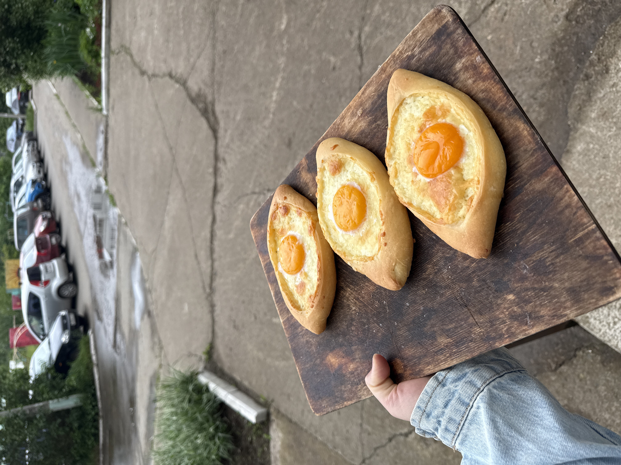 A person, whose left hand and forearm clad in a blue denim sleeve are visible in the lower left, holds a rectangular dark wooden serving board. Positioned diagonally across the board are three boat-shaped Adjaruli Khachapuri, each containing a substantial amount of melted white and lightly browned cheese with a distinct, bright orange egg yolk centrally placed within the cheese filling. The khachapuri appear freshly baked, with a golden-brown crust.
The scene is an outdoor environment in what is identified as Chaikovsky, Russia. The ground beneath and extending into the background is concrete or asphalt, characterized by numerous cracks and visible wet patches, including small puddles, suggesting recent precipitation or damp conditions. A low, white-edged planter box containing green foliage is visible to the left of the person's hand. Further back, a paved area transitions into a parking lane where a series of diverse vehicles are parked in a single file line, receding into the distance. Cars visible include a silver hatchback, a dark sedan, a blue car, and various other colors. Lush green trees and bushes frame the left and right sides of the background, partially obscuring some of the parked vehicles and providing a natural boundary to the paved area. The lighting is diffused, indicating an overcast sky.