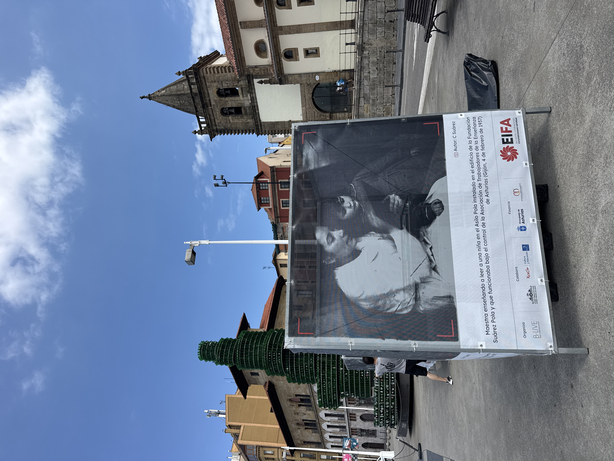 The image shows an outdoor public space in Gijón, Spain, with a large black and white photograph displayed on a stand. The photograph depicts a woman teaching a child to read, identified by text on the stand as being taken in the Asilo Pola, Gijón, on February 4, 1937. In the background, to the left of the photograph, is a spiraling green sculpture made of bottles. Further back, various buildings are visible, including a church with a bell tower. A person in a grey t-shirt and black shorts is standing near the bottle sculpture, facing away from the camera. The sky is blue with scattered clouds. The stand holding the photograph has several logos and text at the bottom.