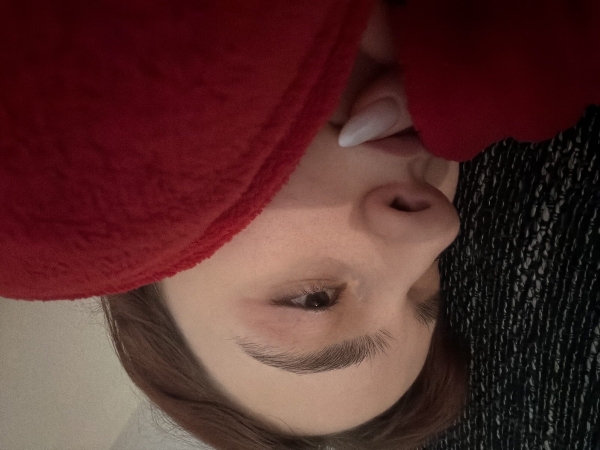 A close-up, angled portrait depicts a person with fair skin and light brown hair. The subject's face is partially obscured by a bright red, soft material on the left side of the frame. The person's right eye, a dark brown, is visible, as well as a defined eyebrow. The nose is prominent, and the mouth is partially seen, revealing a white fingernail. The background features a dark fabric or material with a textured pattern. The location in Strasbourg, France, is unknown.