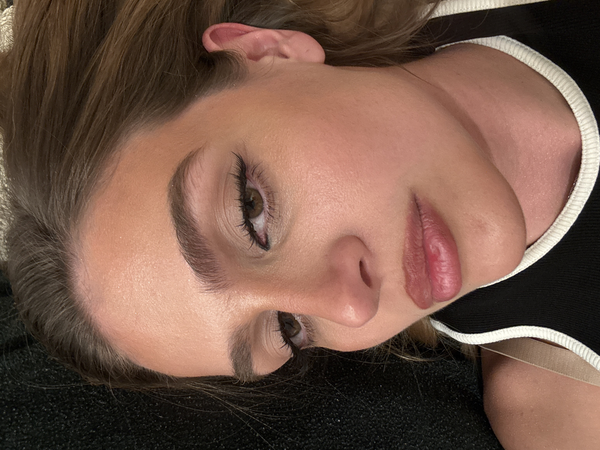 A close-up portrait shows a person, likely female, lying down with their head angled to the right, looking downwards and to the left of the frame. Her light to medium brown, wavy hair frames her face. Her skin appears smooth with a subtle sheen on the forehead, cheekbones, and nose, possibly due to natural oils or makeup. She has prominent dark eyelashes, dark brown eyes, and well-defined eyebrows. Her full, pinkish-red lips are slightly parted, revealing no teeth.
She is wearing a black top with a distinctive white ribbed trim along the neckline, and a glimpse of a light-colored bra strap is visible underneath. The person is resting on a dark, textured surface, possibly a blanket or upholstered furniture, which occupies the lower left portion of the image. The lighting is soft and even, suggesting an indoor setting. Her expression is relaxed and neutral. No text is visible in the image.