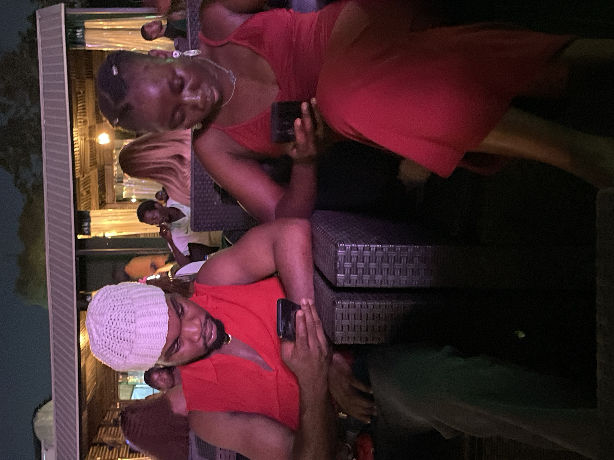 The image depicts a scene at night, likely outdoors in the city of Abuja, Nigeria. Two individuals are seated in outdoor furniture, possibly at a restaurant or bar, as suggested by the ambient lighting and background structures.
The person on the left is a man wearing a red sleeveless shirt and a white knitted beanie. He is looking down at his mobile phone, which he is holding in his right hand. His left arm rests on the furniture beside him.
The person on the right is a woman wearing a red dress with spaghetti straps. She is also looking down at her mobile phone, which she is holding in her right hand. Her left arm is bent, resting near her lap.
In the background, other people are visible, some are seated and some are standing. The environment appears to be a covered outdoor area with lighting fixtures. The overall impression is a relaxed, social setting at night.