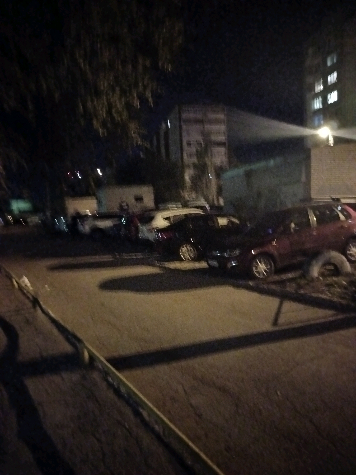 An outdoor nighttime scene in the city of Astrakhan, Russia, shows parked cars on a street in front of a brick building. A beam of light, likely from a searchlight or floodlight, is directed from the brick building towards the sky, illuminating a portion of the darkness. Several cars are visible, including a red one on the right side of the frame and a black one to its left. Some trees are also present, casting shadows on the asphalt. The image appears to be taken from a low angle.