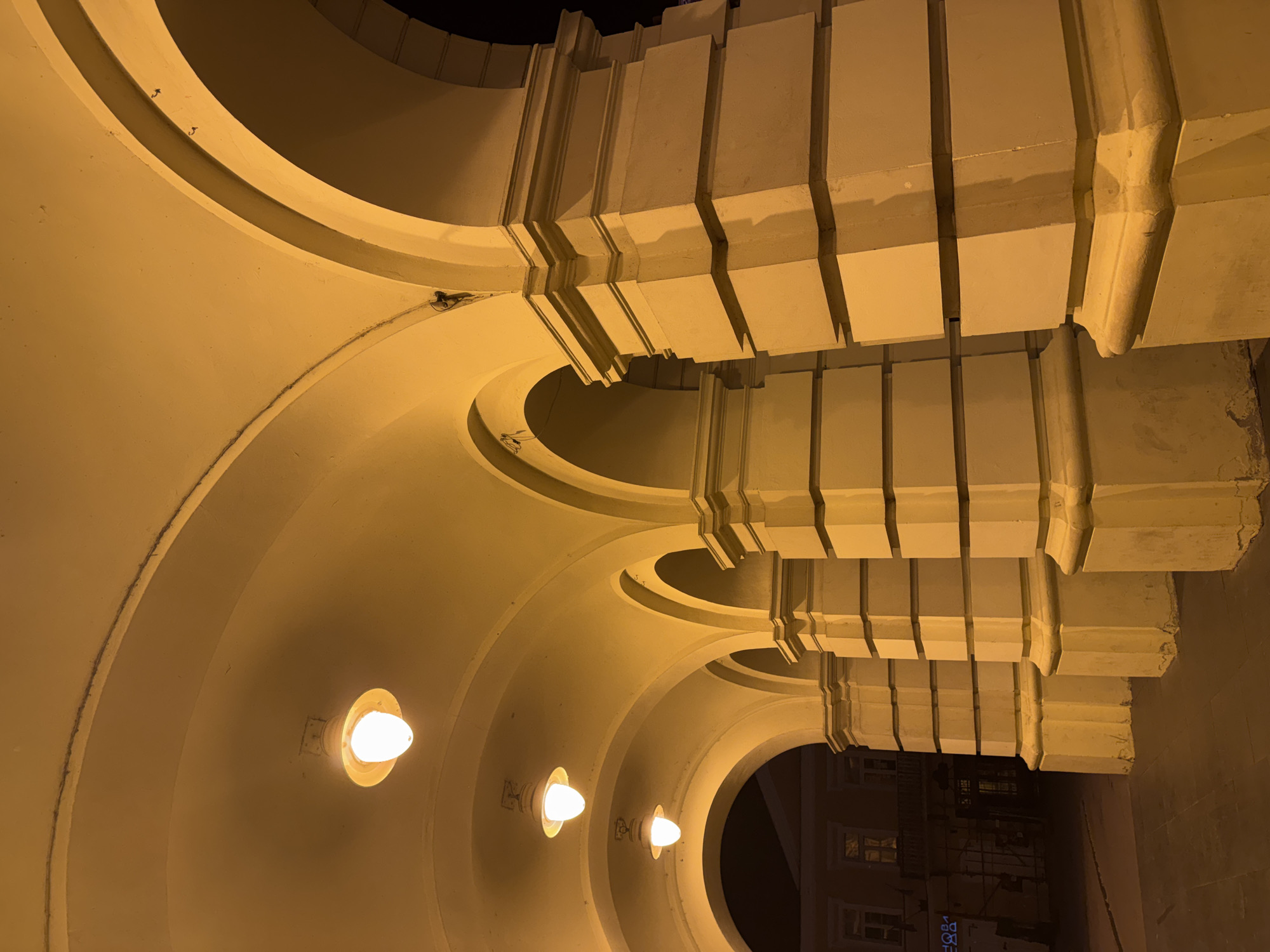 The image depicts a series of illuminated arches and columns, likely part of a historic building or public walkway in Novi Sad, Serbia. The architecture features classical elements with segmented columns supporting rounded archways. Several pendant lights, emitting a warm yellow glow, are visible along the curved ceilings of the arches, illuminating the scene at night. The foreground shows a paved ground surface, possibly a sidewalk or street, which is also lit by the overhead lights and ambient street lighting. In the background, through the arches, glimpses of a building with windows and balconies can be seen, suggesting an urban setting. The overall impression is one of a well-maintained, dimly lit architectural space.