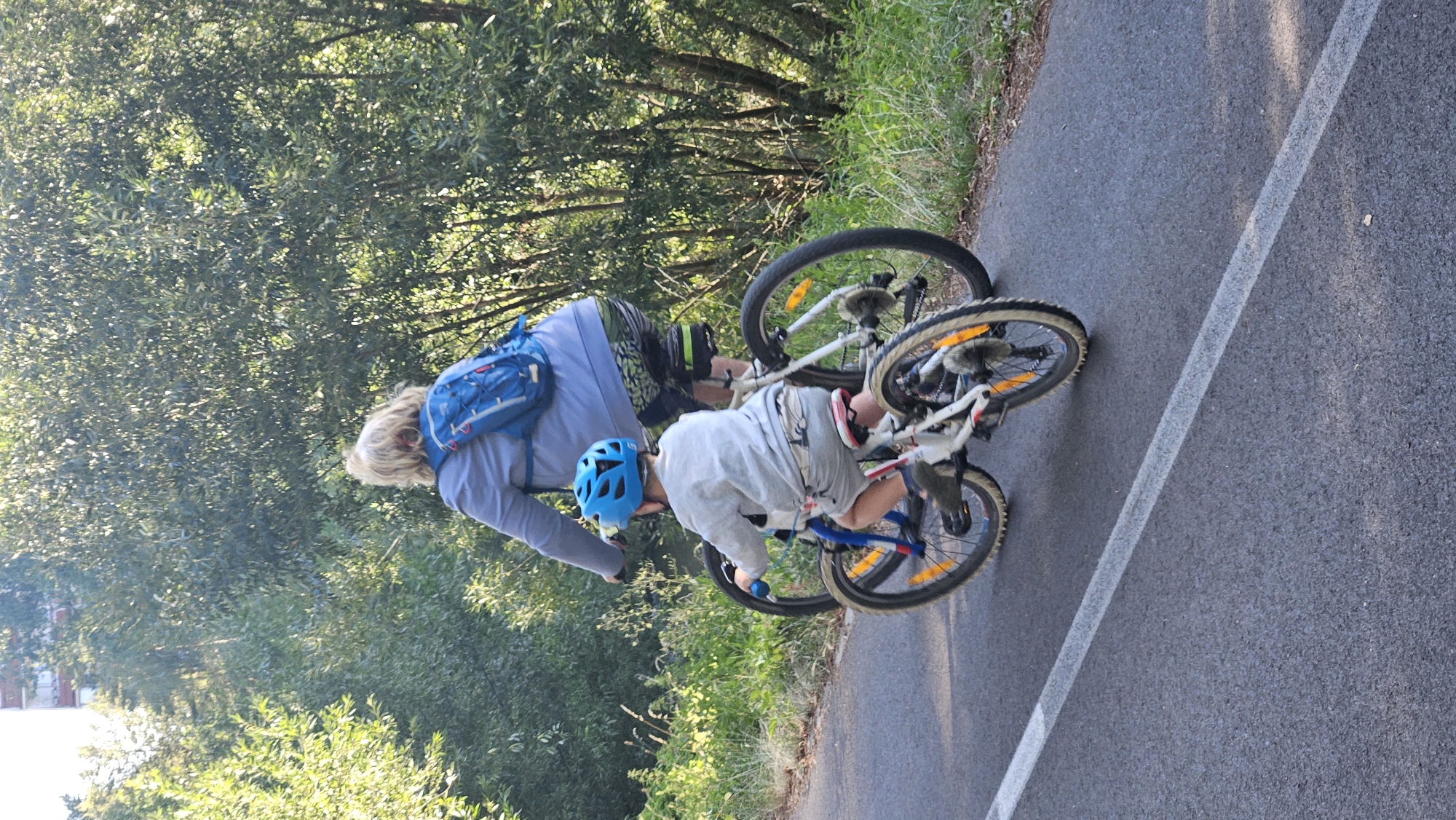 The image shows two individuals cycling on a paved path. The person in the foreground, appearing to be a child, is wearing a blue helmet and a light-colored long-sleeved shirt and shorts, riding a white bicycle. The person in the background, appearing to be an adult, is wearing a blue backpack and cycling behind the child. Both are on a paved path bordered by greenery, suggesting an outdoor recreational area. The context of Prešov, Slovakia, is not visually discernible from the image itself.