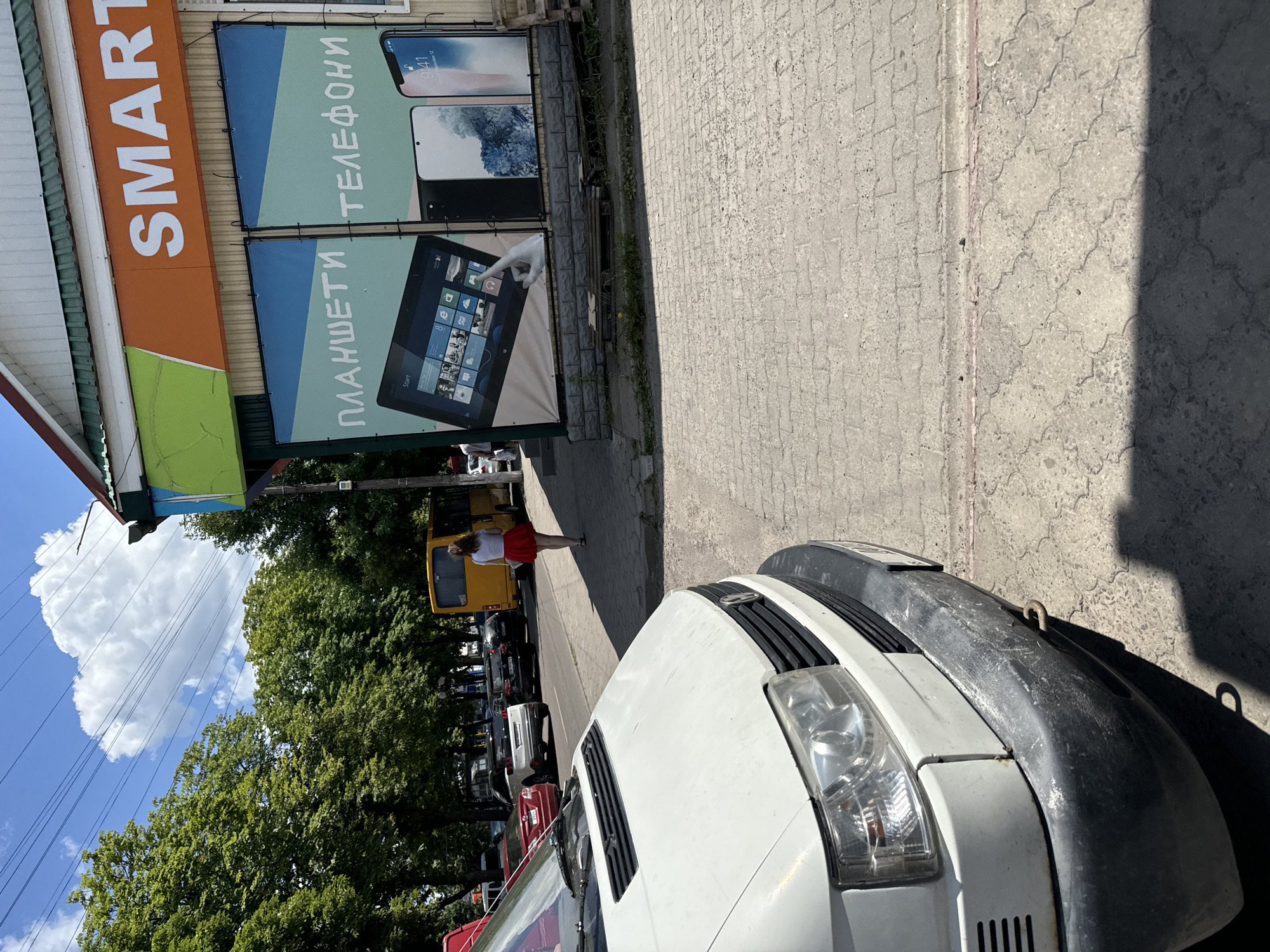 The image depicts a street scene in Sarny, Ukraine. The setting is an outdoor commercial area with a building on the left displaying a large orange sign with white lettering that reads "SMART". Below this sign are two large banners advertising mobile devices. The top banner shows two smartphones and has Cyrillic text that translates to "TELEPHONES". The bottom banner features a tablet displaying a Windows interface and has Cyrillic text that translates to "TABLETS".
In the foreground, the front of a white van is visible, with a dark gray bumper. The van is parked on a paved surface. A woman in a red skirt and white top is walking away from the camera on the sidewalk. Behind her, a yellow van is parked, and further down the street, several other vehicles are lined up. Lush green trees border the street, and a bright blue sky with fluffy white clouds is visible above. The lighting suggests it is daytime, likely mid-morning or afternoon, with clear weather. The overall scene conveys a casual, everyday atmosphere of a commercial street.