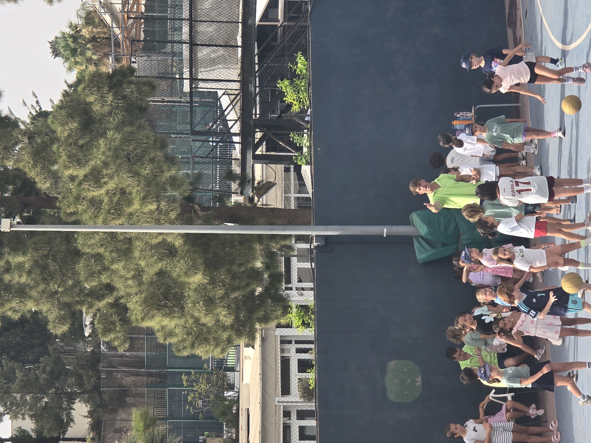 The image shows a group of children and an adult male on an outdoor court, possibly a tennis or basketball court, in Málaga, Spain. The adult, wearing a bright green polo shirt, appears to be instructing or leading the children. Several children are in athletic attire, some are holding or playing with a basketball. The background reveals a dark blue wall, fences, trees, and buildings, suggesting an urban or park setting. One child in the foreground is wearing a jersey with the number 17 and the name "CARLA". The lighting indicates it is daytime.