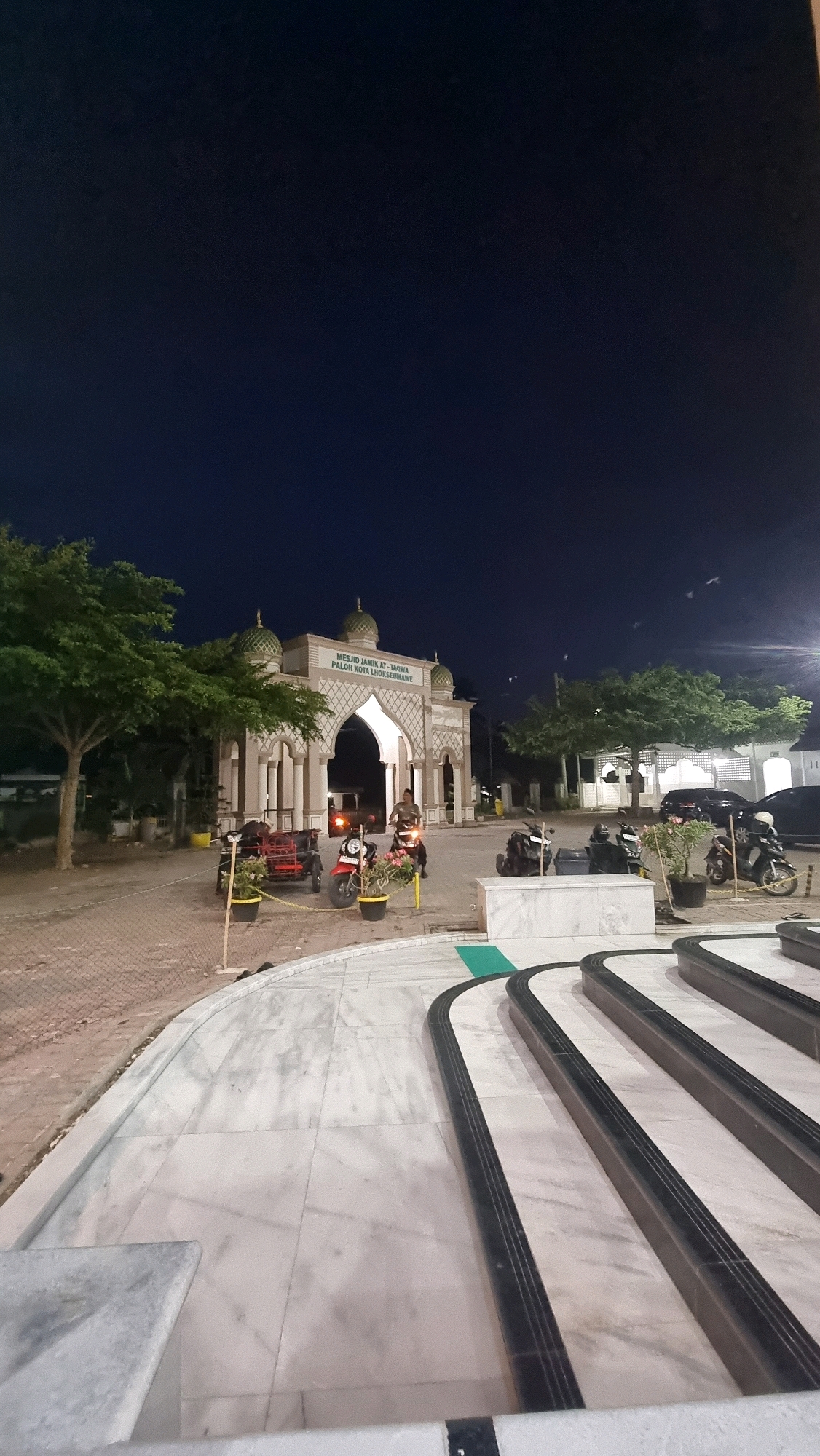The image depicts the entrance of Masjid Jamik At-Ta'qwa in Paloh, Lhokseumawe City, Indonesia, at night. The mosque features a distinct architectural style with multiple domes and an arched gateway. In front of the mosque, there are several parked motorcycles, a red ATV, and a few people. One person is seated on a motorcycle near the entrance, while another appears to be standing. The surrounding area includes trees, paved pathways, and some decorative marble structures in the foreground. The sky is dark, indicating it is nighttime, and artificial lighting illuminates the scene.