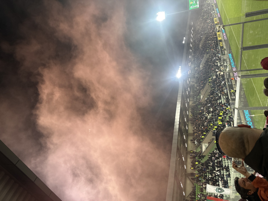 The image displays a nighttime scene inside a sports stadium located in Razgrad, Bulgaria. The upper portion of the stadium is filled with dense, reddish-pink smoke, obscuring the overhead sky. Below the smoke, multiple tiers of spectator seating are populated by numerous individuals, mostly dressed in dark attire. Scattered among the crowd are persons wearing yellow high-visibility vests. In the immediate foreground, the back of a bald head is visible from a spectator's perspective, while to the left, another individual holds a smartphone, actively capturing a photograph or video. Bright floodlights, emitting strong beams, illuminate the stadium from the right side, cutting through the smoke. A green football pitch is visible at the bottom of the frame, bordered by advertising hoardings featuring Cyrillic text, including "Еfbet" and "СТАТИСТИКА В РЕАЛНО ВРЕМЕ". A digital scoreboard on the far right displays the numbers "19:42" and "3". A white and green flag bearing a crest is visible within the lower-left section of the stands. A dark structural element, likely part of the stadium's roof or canopy, is partially visible in the upper-left corner.