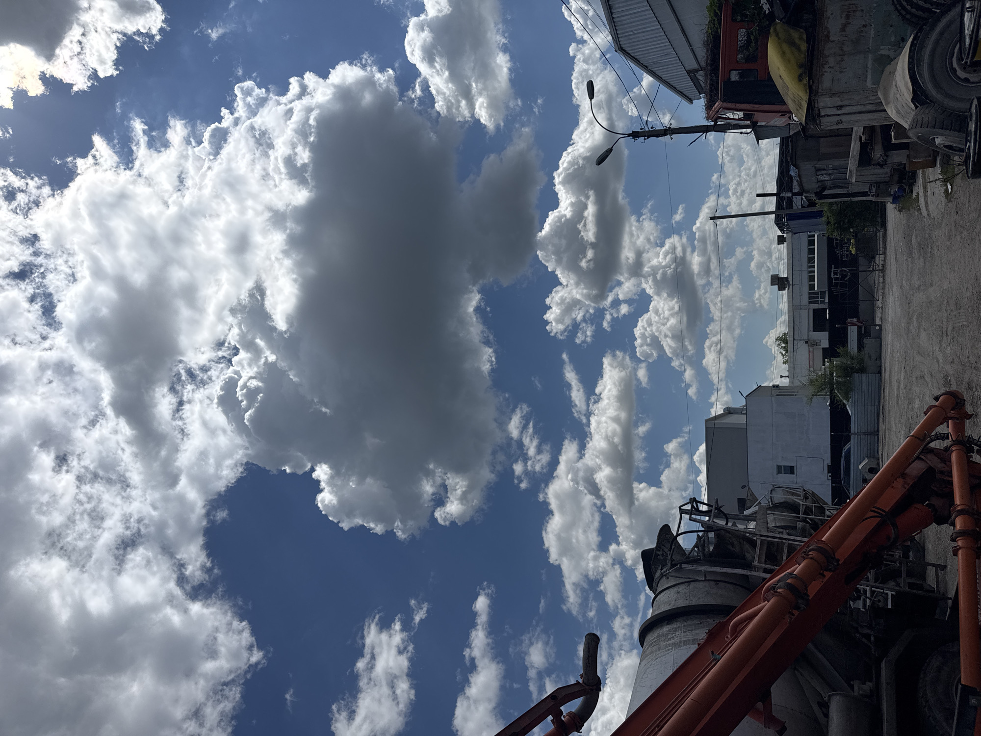 The image shows a construction site under a partly cloudy sky. In the foreground, a concrete mixer truck with an orange boom is visible on the left. The background features various industrial buildings, a streetlamp, power lines, and a red truck. The ground is dirt and gravel. The scene appears to be in an urban industrial area.
