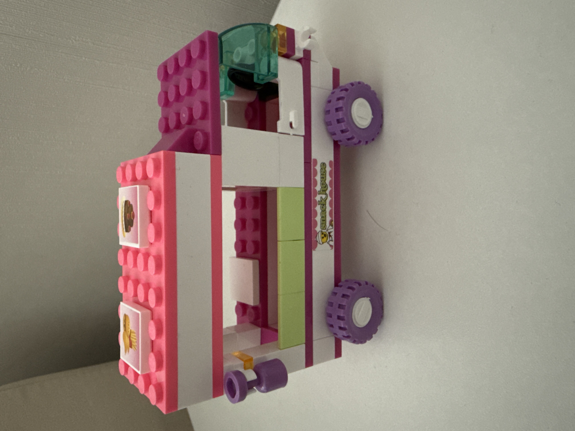 A Lego Friends "Snack House" truck, primarily pink and white, is positioned against a white or light grey surface. The truck has purple wheels. The scene is indoors. The location in the city of Верхнеказымский, Russia, is not identifiable from the image.