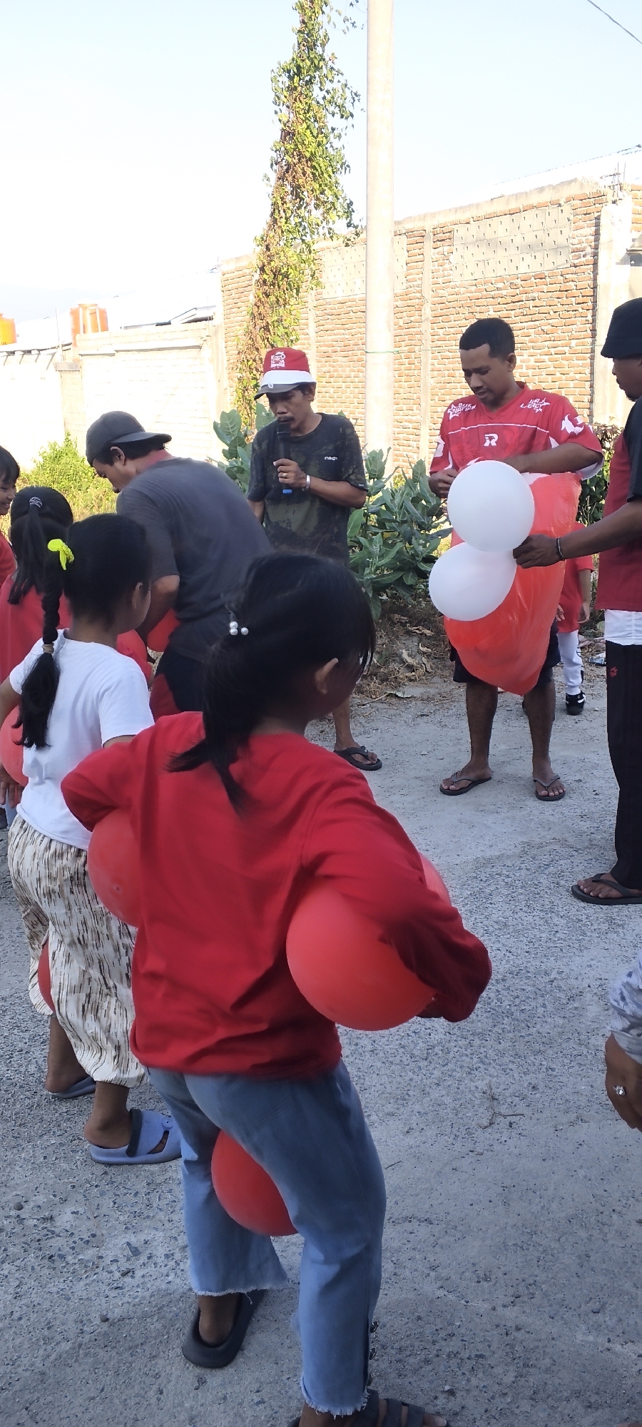 In Tawanjuka, Indonesia, a group of individuals, predominantly children wearing red shirts, are gathered outdoors on a gravel surface. A man in a red shirt and cap holds a microphone, speaking to the group, while another man in a red shirt stands nearby with a large red bag containing two white balloons. Two children in the foreground are positioned to hold red balloons between their bodies. The scene is set against a backdrop of a brick wall and foliage under a clear sky.