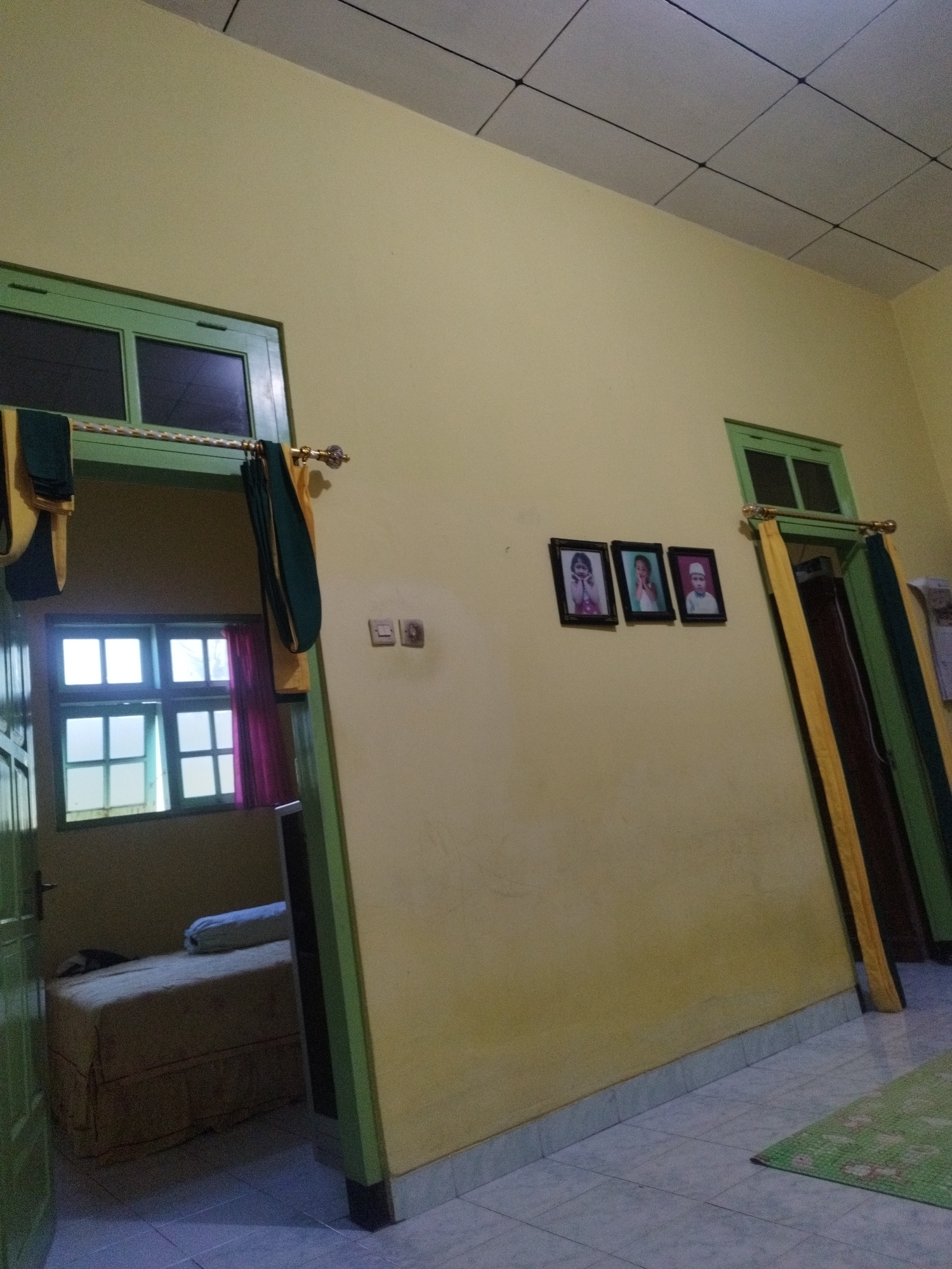 The image depicts an indoor scene, likely a bedroom or living area in a residential dwelling. The walls are painted a pale yellow, and the ceiling is composed of white acoustic tiles with black grid lines. Natural light filters in from two windows, one visible through an open doorway on the left and another to the right. The window on the left has a green frame and is divided into multiple panes. Yellow and dark green curtains hang from a rod above this doorway.

To the right, another window with a green frame is partially obscured by yellow and dark green curtains. Three framed photographs are displayed on the yellow wall between the two windows. The photographs appear to be of children. On the wall next to the right window, there are two light switches.

The doorway on the left opens into a room containing a bed with a patterned cover and a bolster pillow. The bed is positioned on the left side of this room, with a dark brown object, possibly a piece of furniture, to its right. The floor throughout the visible area is tiled with light blue and gray squares. In the lower right corner of the image, a portion of a green rug with a floral pattern is visible on the floor.

Based on the interior design elements, such as the window frames, curtains, and the general aesthetic, this dwelling is consistent with residential architecture found in Sumenep, a city located on the island of Madura in East Java, Indonesia. The simple furnishings and the presence of family photographs suggest a domestic setting typical of middle-income households in the region.
