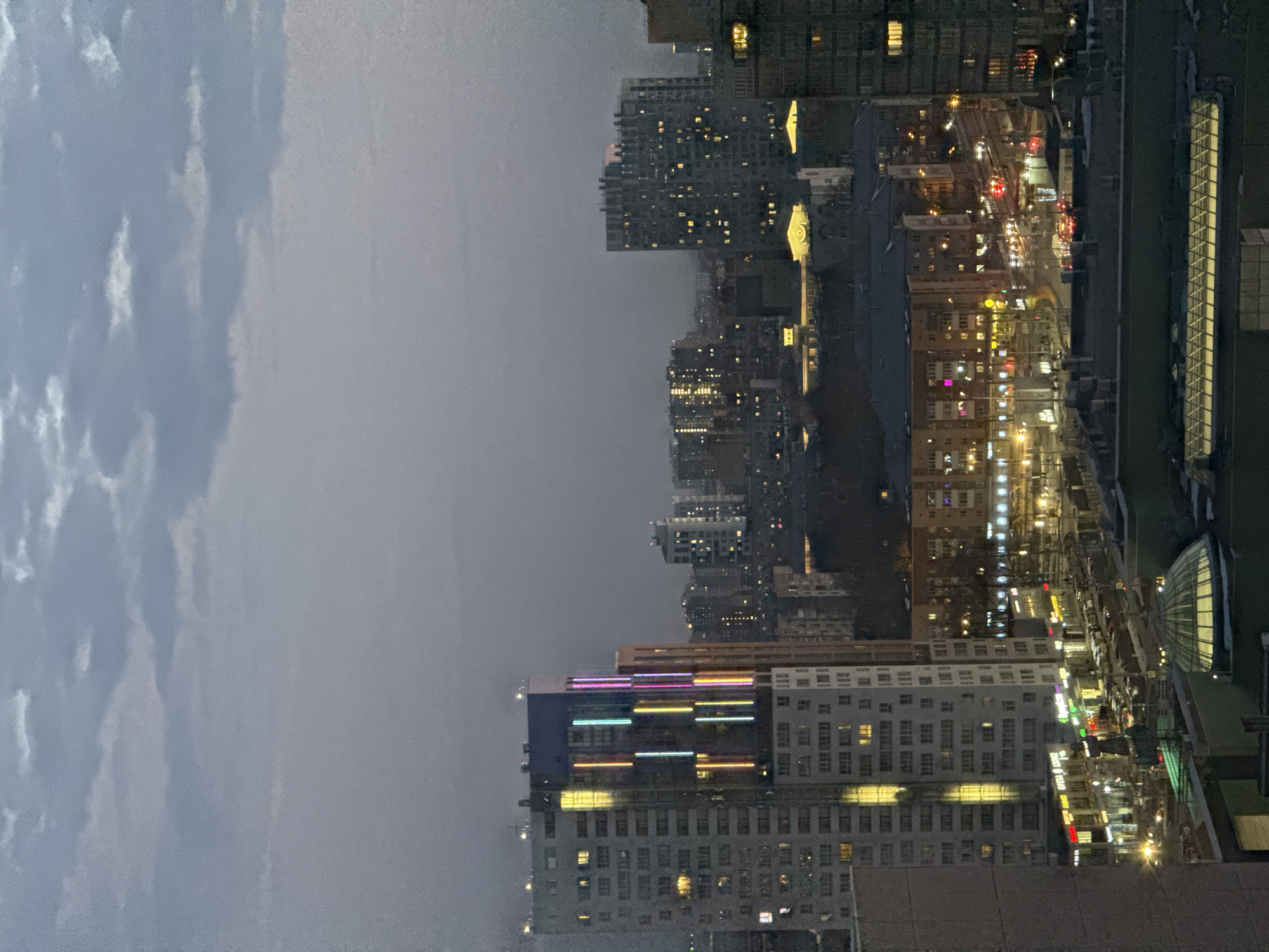 The image depicts a nighttime cityscape taken from an elevated position, possibly a high-rise building. The left side of the image shows a cloudy sky, transitioning into the city view on the right. The city is densely populated with buildings of varying heights, illuminated by streetlights and lights emanating from within the structures. There is evidence of vehicular traffic on the streets. The overall atmosphere suggests a damp or overcast evening. The image does not provide enough specific detail to identify Perm, Russia.